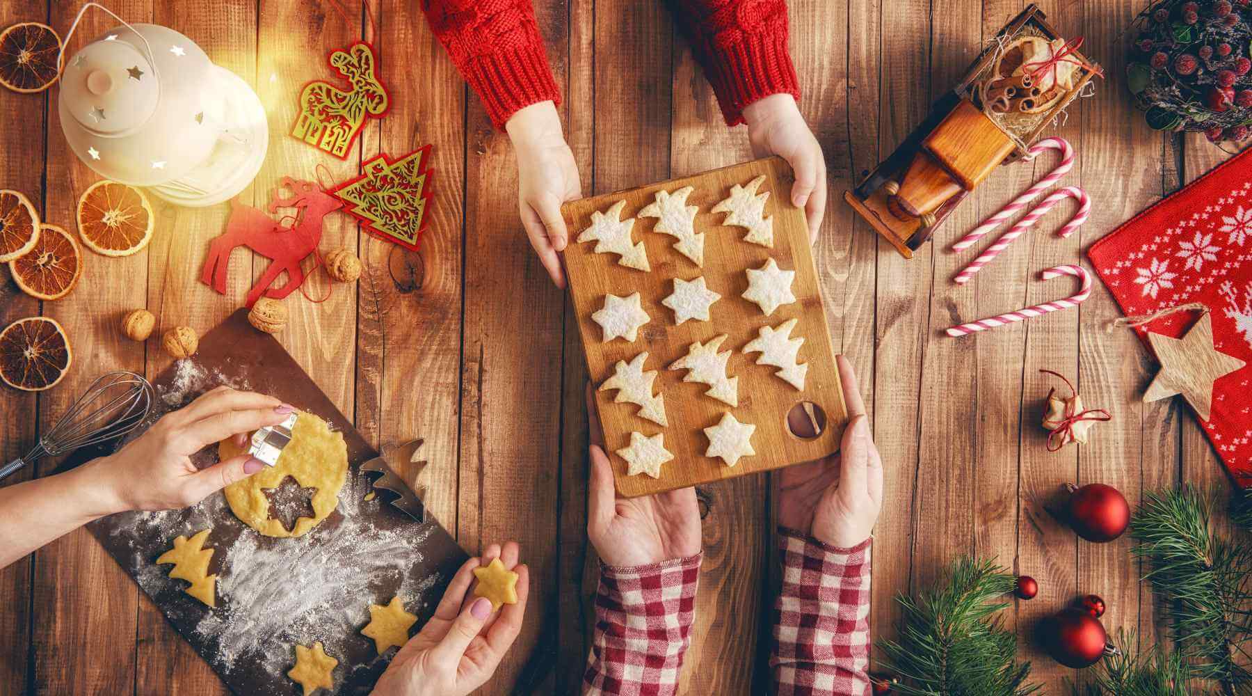 Family members baking Christmas cookies together, with hands holding a wooden tray filled with star and tree-shaped cookies, festive decorations like dried orange slices, and holiday ornaments scattered on a rustic wooden table.