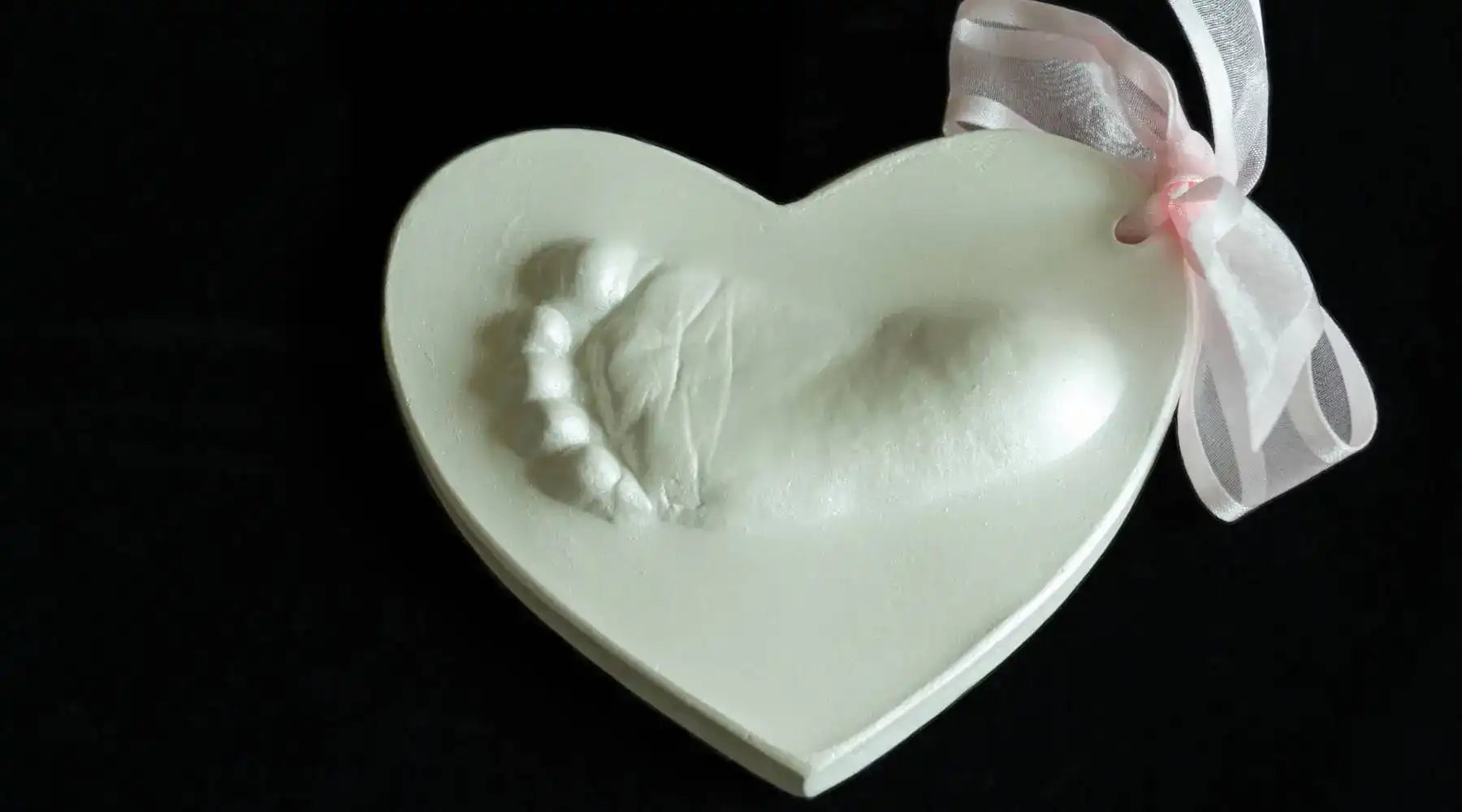 Heart-shaped white plaque with baby footprint impression and pink ribbon, perfect keepsake for newborns.