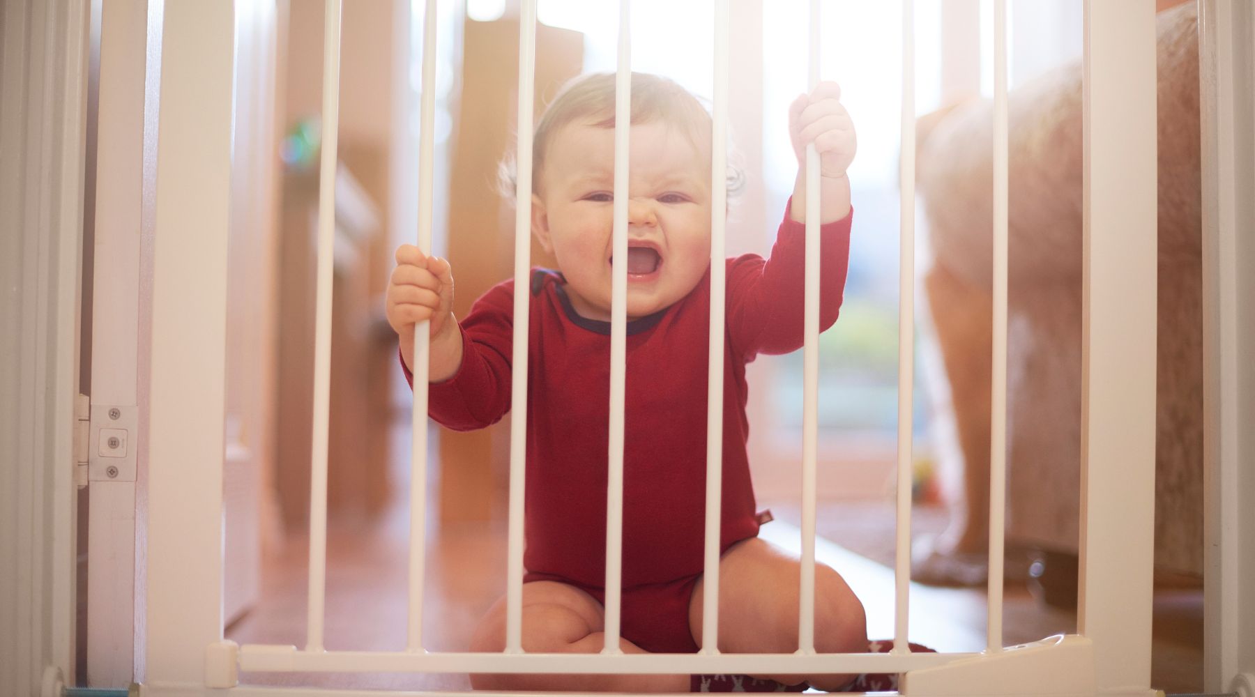 Baby behind a white baby gate, appearing upset while reaching out.