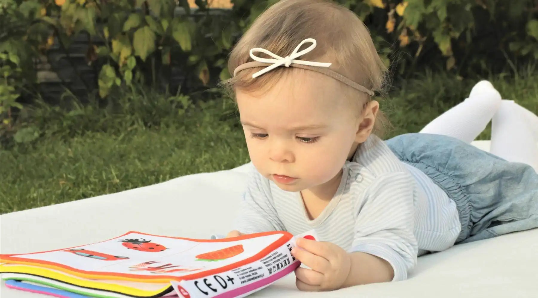 Baby girl lies outdoors reading soft book, exploring textures and images to boost early brain development.