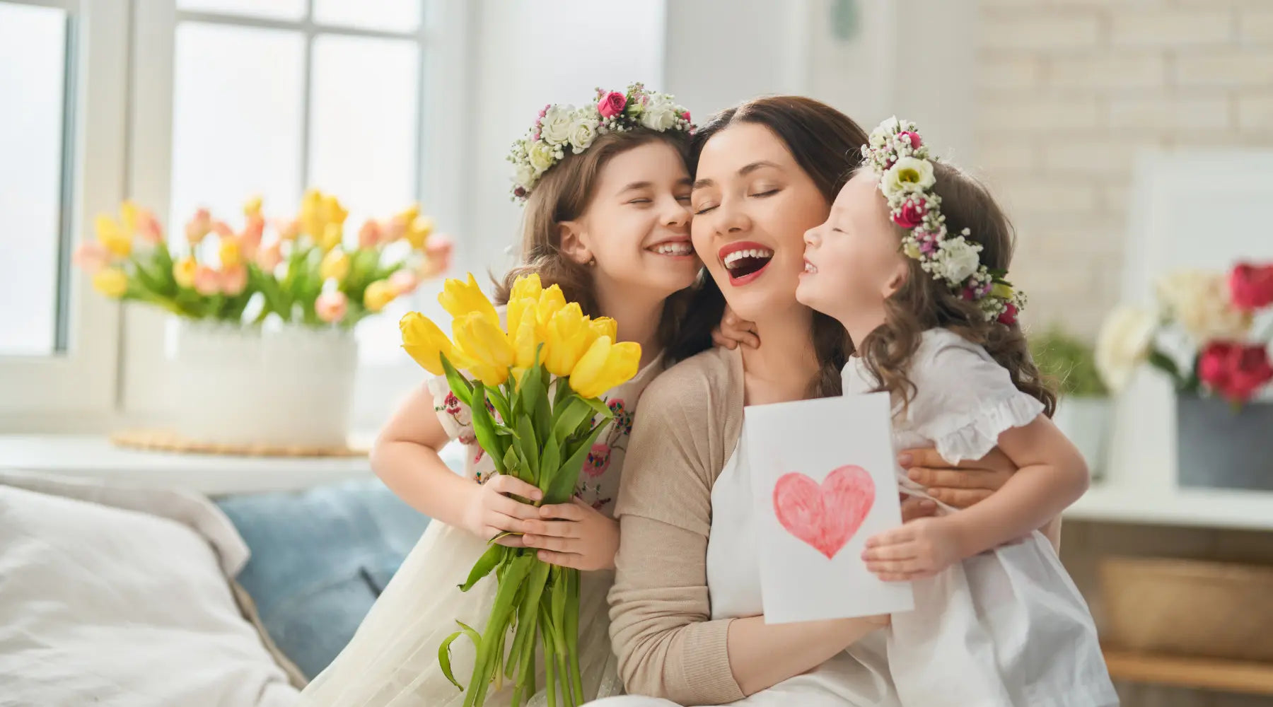 A joyful mother sits with her two daughters, who are hugging her and presenting her with a bouquet of yellow tulips and a handmade card featuring a red heart. The girls wear white dresses and floral crowns, creating a warm and loving moment.