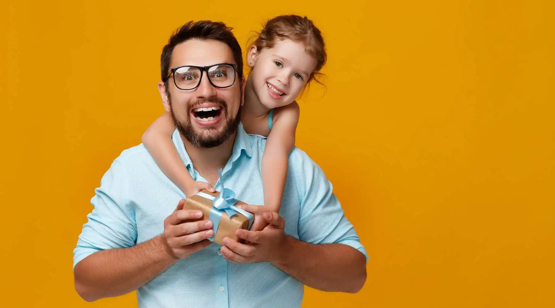 Man in glasses holding gift with blue ribbon as young girl hugs him from behind, yellow backdrop