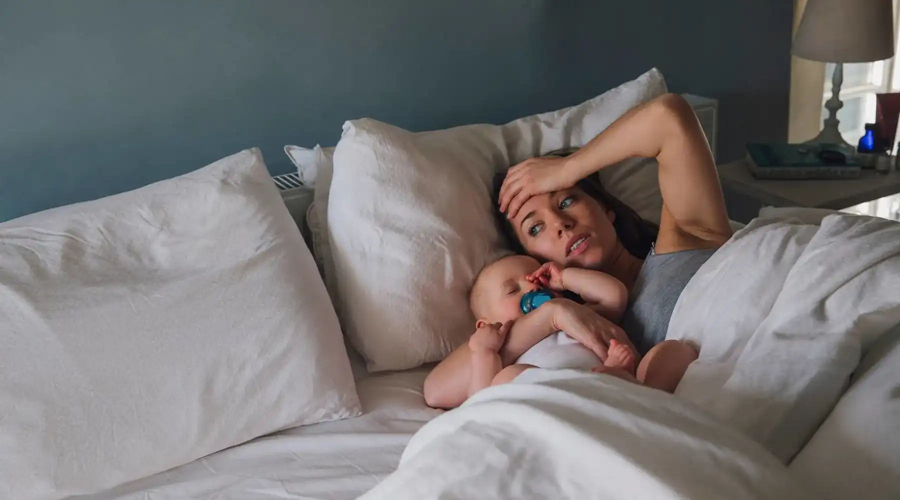 Mom with hand on forehead looks down while feeding baby, appearing stressed in cozy nursery.