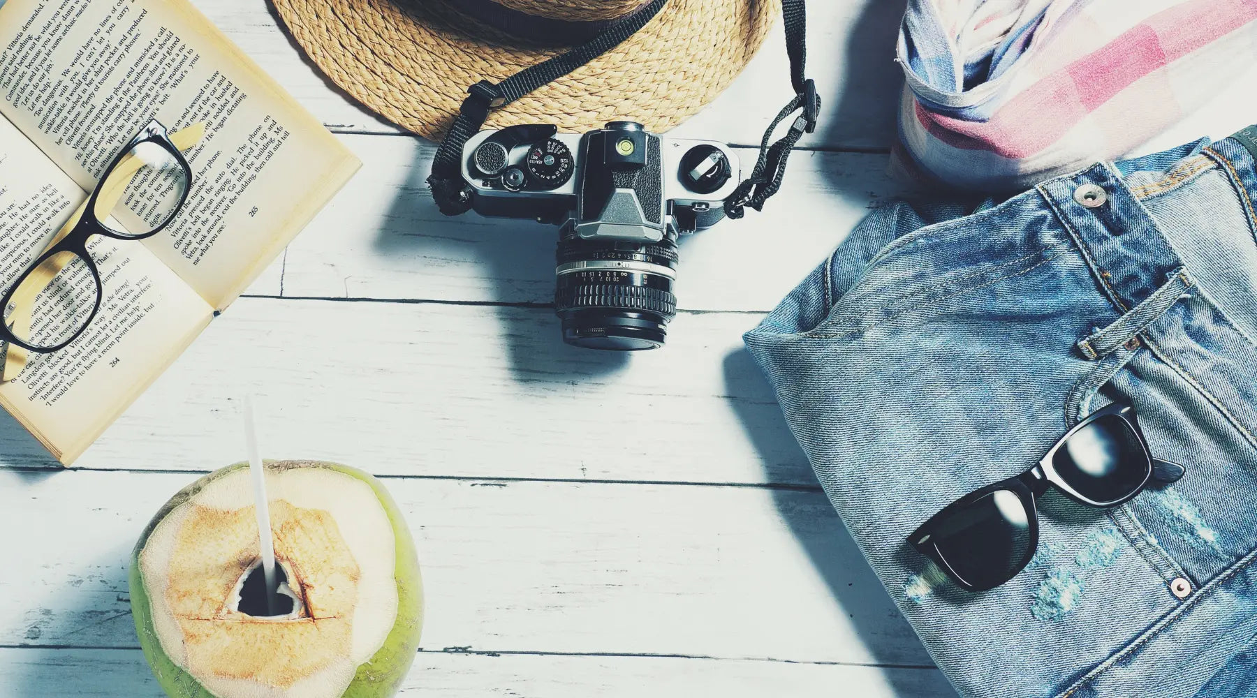 A collection of summer travel essentials arranged on a white wooden surface, including a straw hat, vintage camera, denim shorts, sunglasses, a book with reading glasses, and a fresh coconut with a straw.