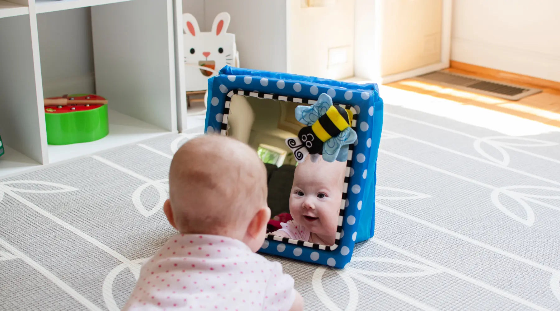 A baby engaging in tummy time while looking at their reflection in a soft, blue polka-dotted baby mirror with a plush bee decoration, promoting sensory development.