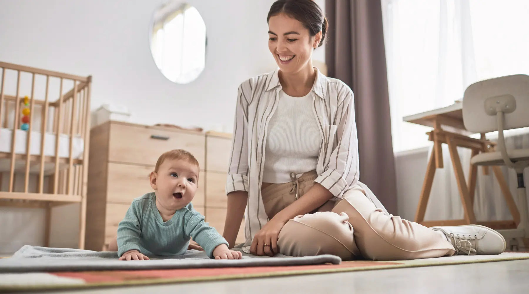 A smiling mother watches her baby engage in tummy time on a soft mat in a well-lit, modern nursery, fostering early development and connection.
