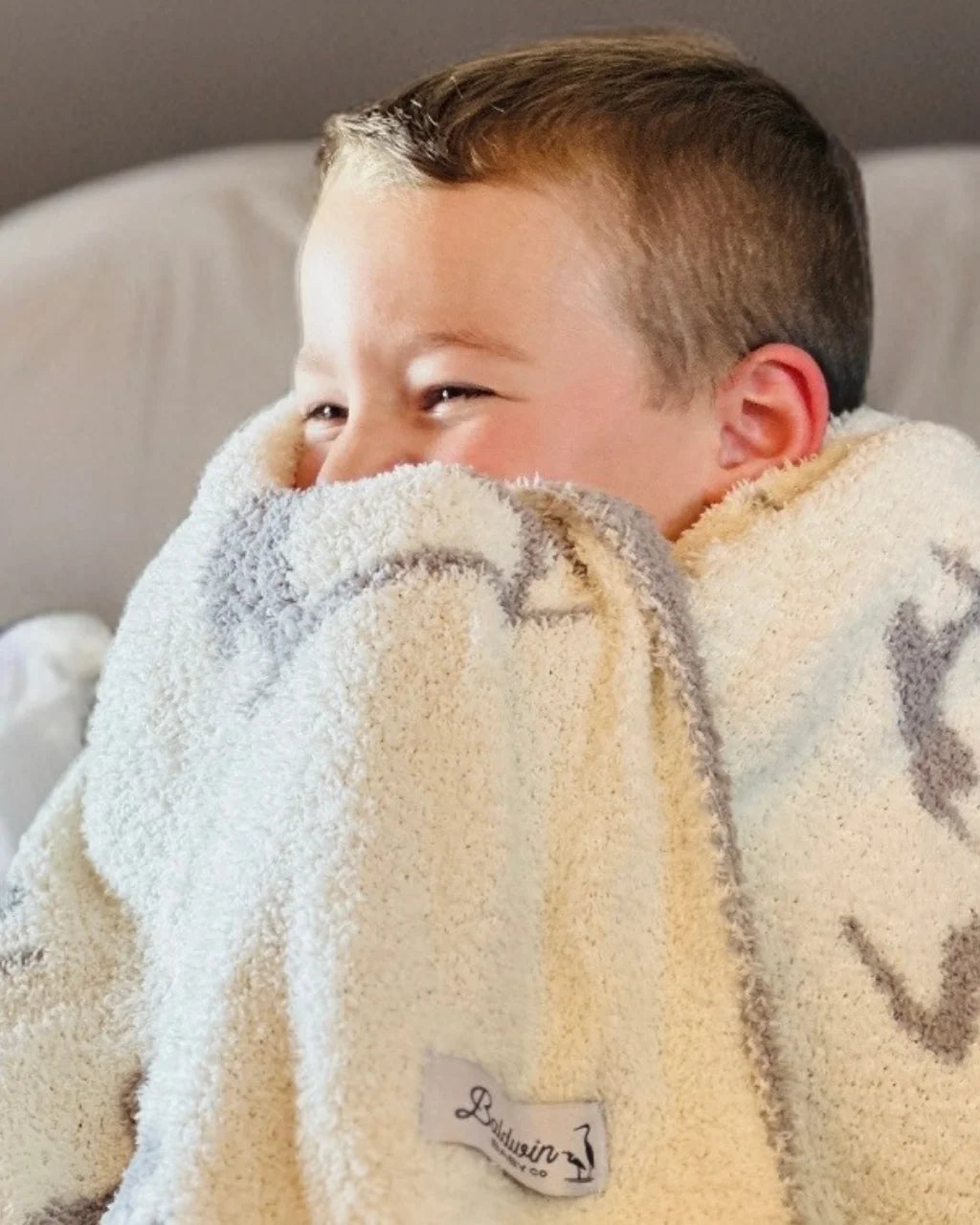 Young boy laughing and snuggled into a creamy white with a gray mother and baby heron silhouette pattern blanket