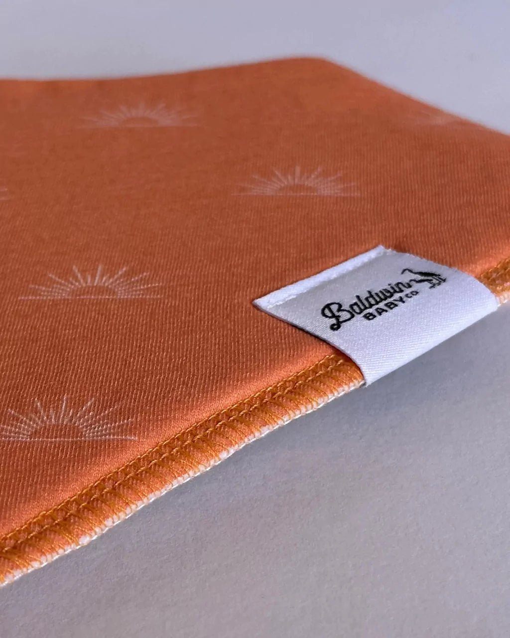 Close up of orange with white sunset pattern bandana bib with a white Baldwin Baby Company logo tag