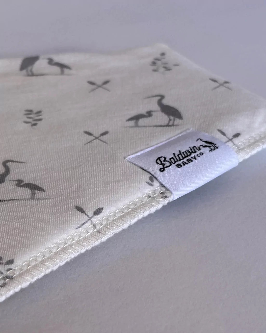Close up of creamy white with Baldwin Baby Co's signature heron pattern bandana bib with a white Baldwin Baby Company logo tag
