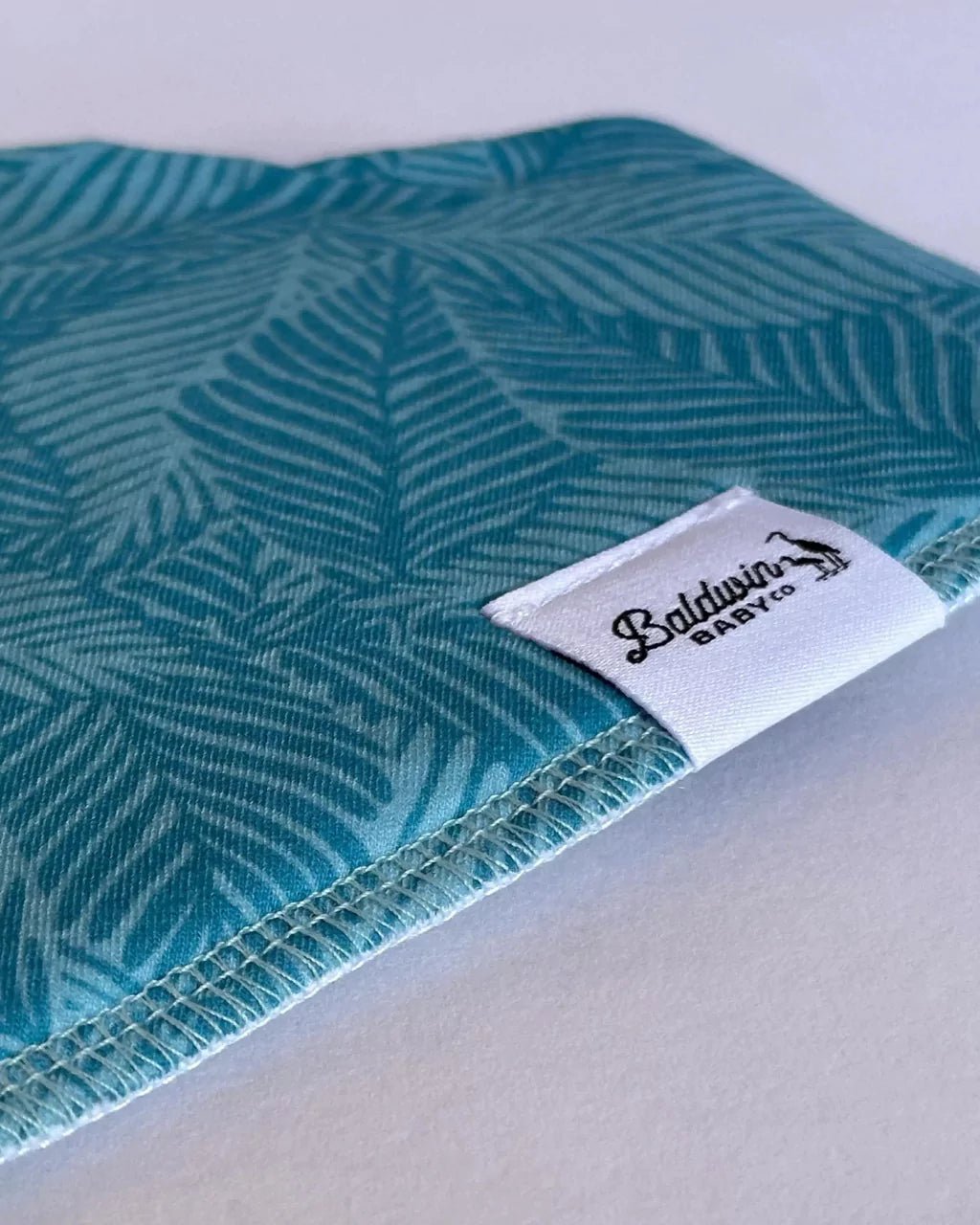 Close up of green monstera pattern bandana bib with a white Baldwin Baby Company logo tag