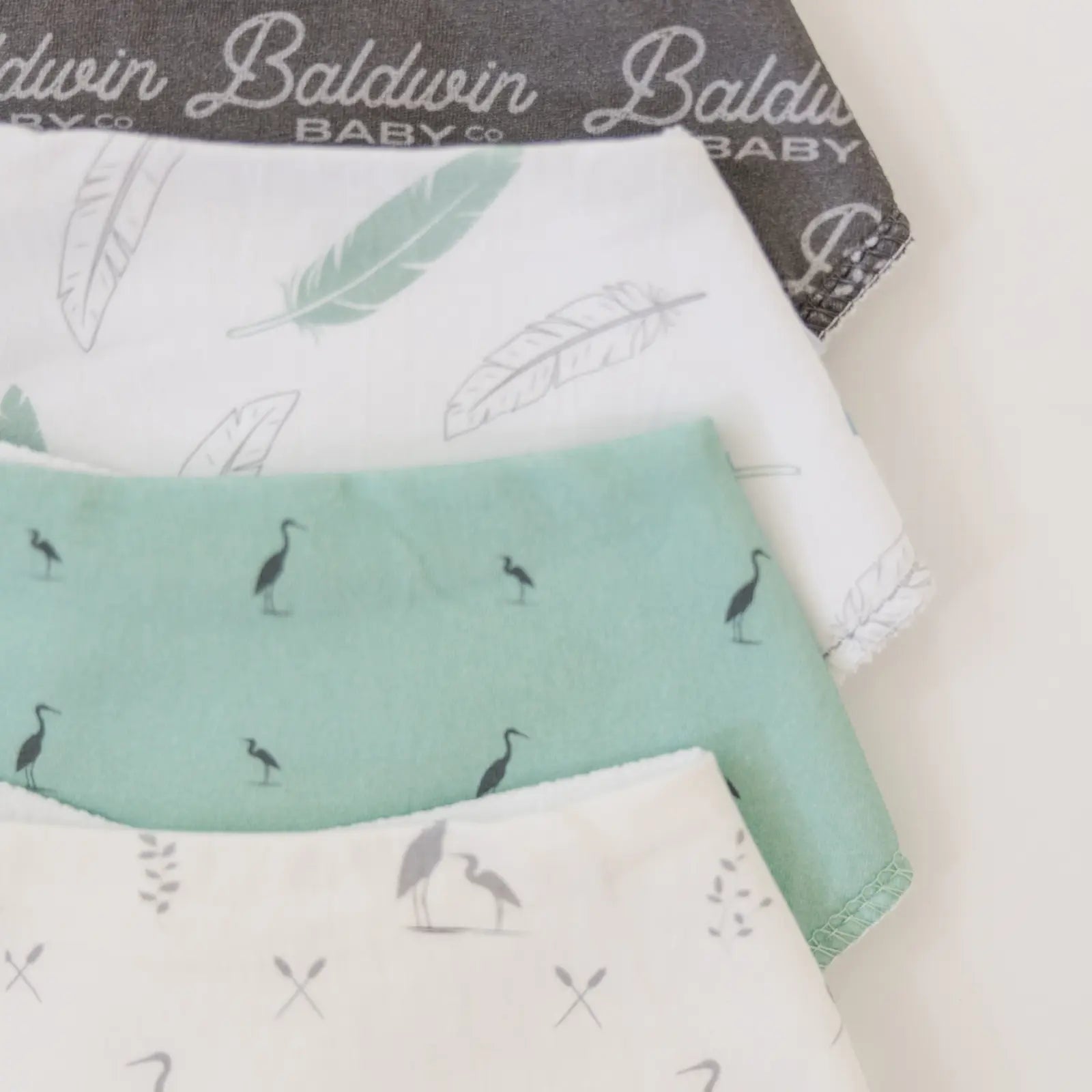 Close-up of four Baldwin Baby Co. bandana bibs with feather and heron designs in various colors.