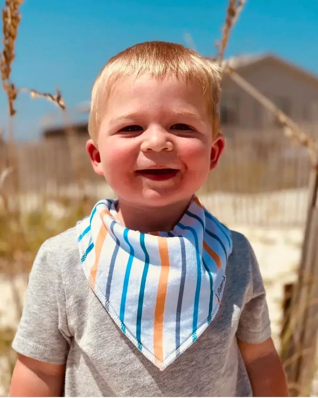 Little boy on the beach while wearing a white, blue, gray, and orange striped pattern bandana bib