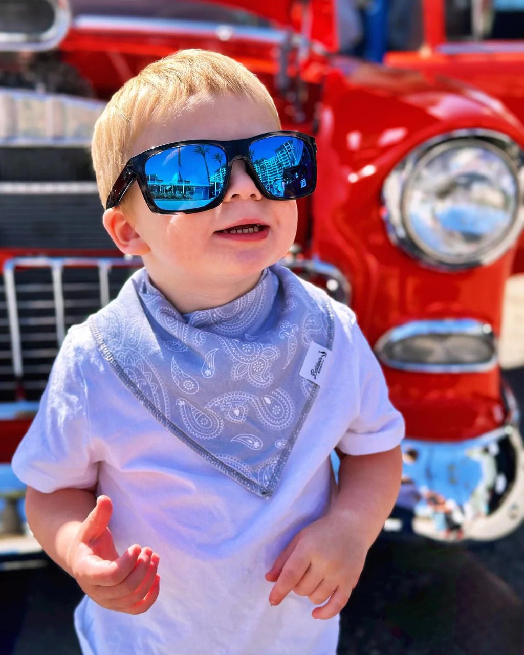 Toddler in front of red car wearing sunglasses and gray paisley bandana bib by Baldwin Baby Co