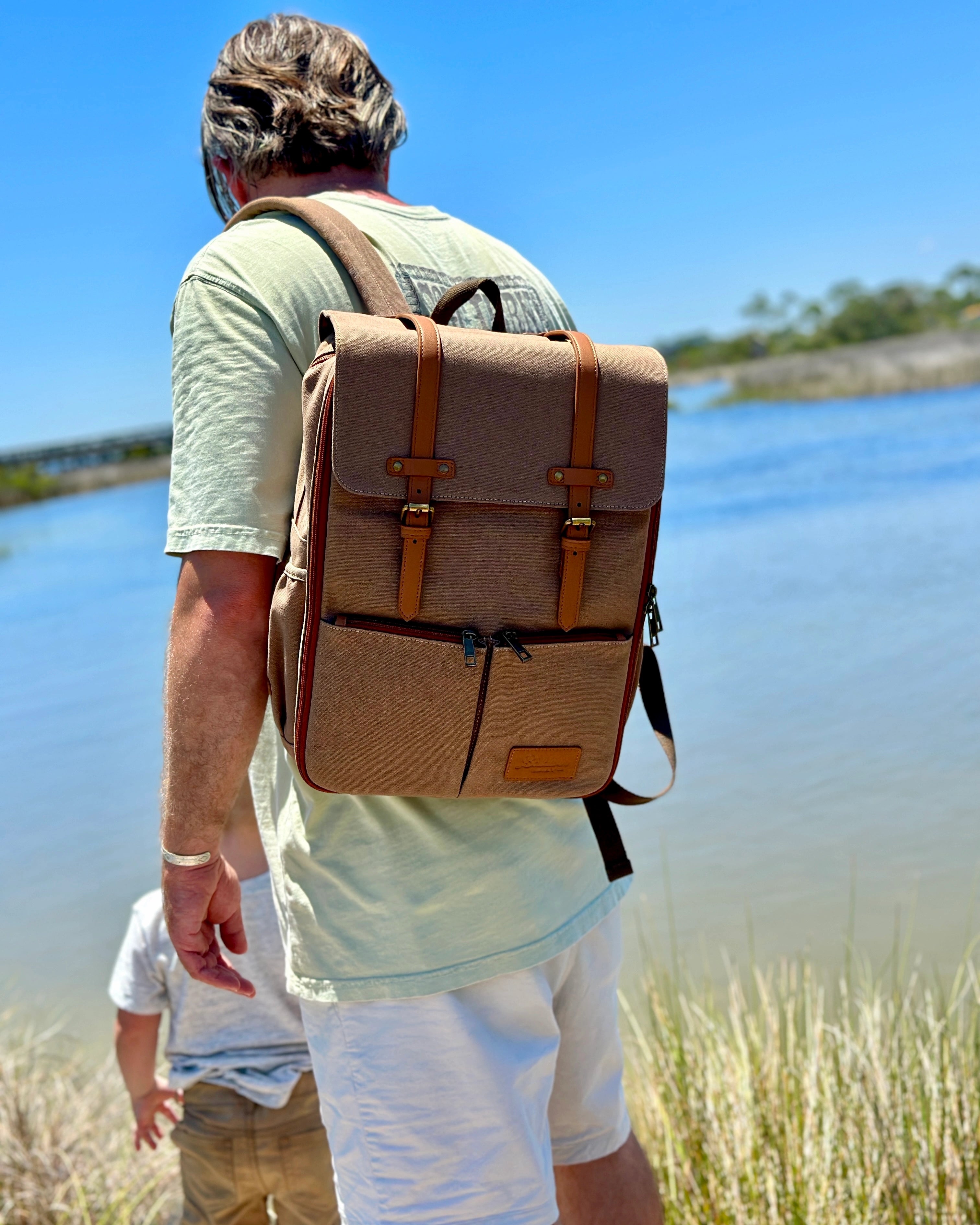Man wearing canvas diaper backpack while watching toddler by the water