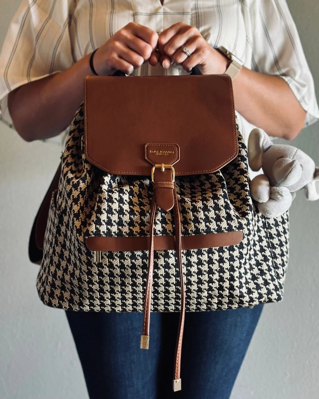 Front view of houndstooth diaper backpack with brown flap, held by woman in casual outfit