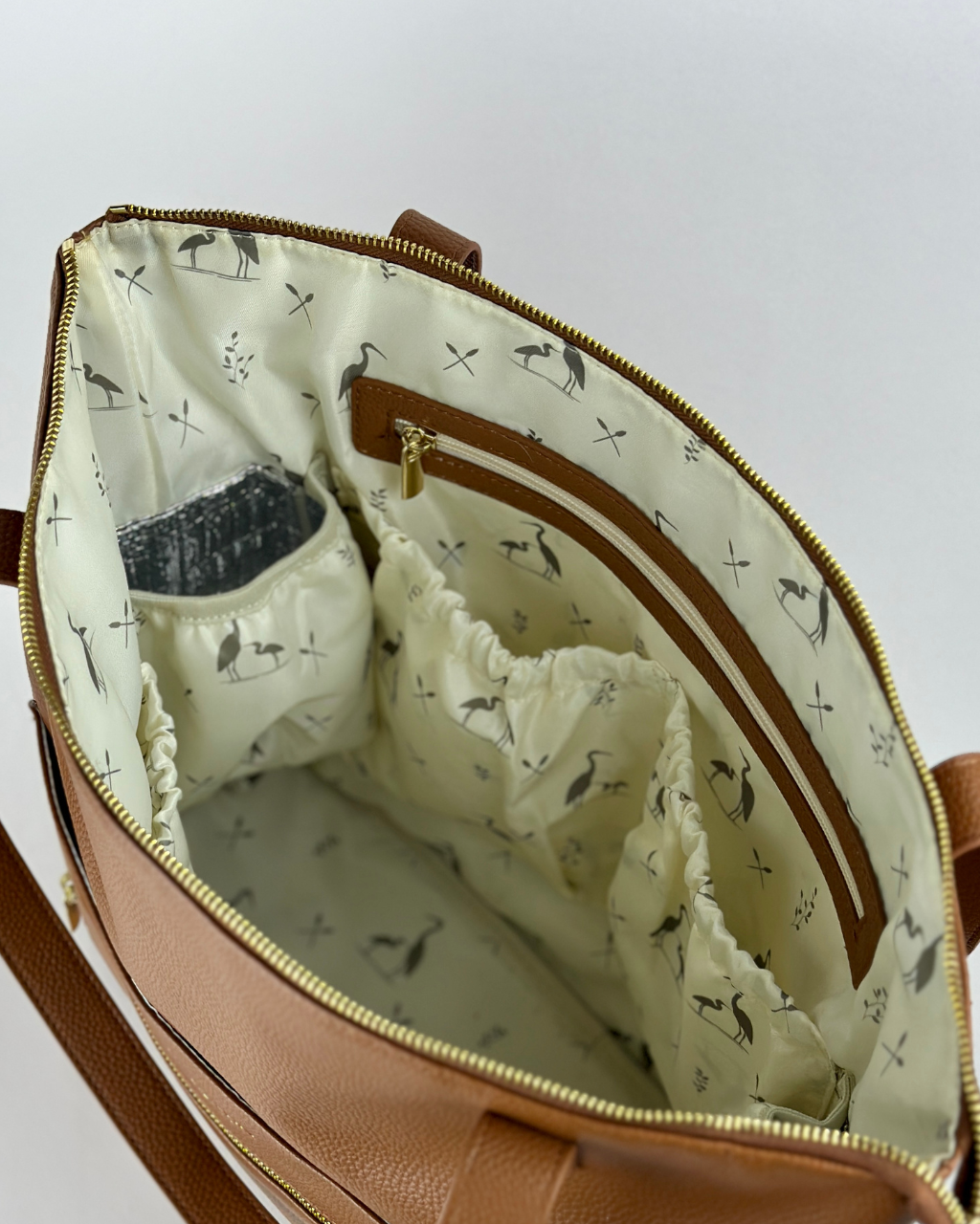 nterior view of the Cognac Lillian diaper bag, featuring a spacious design with multiple elastic and zippered pockets, lined with elegant heron-themed fabric for stylish organization.