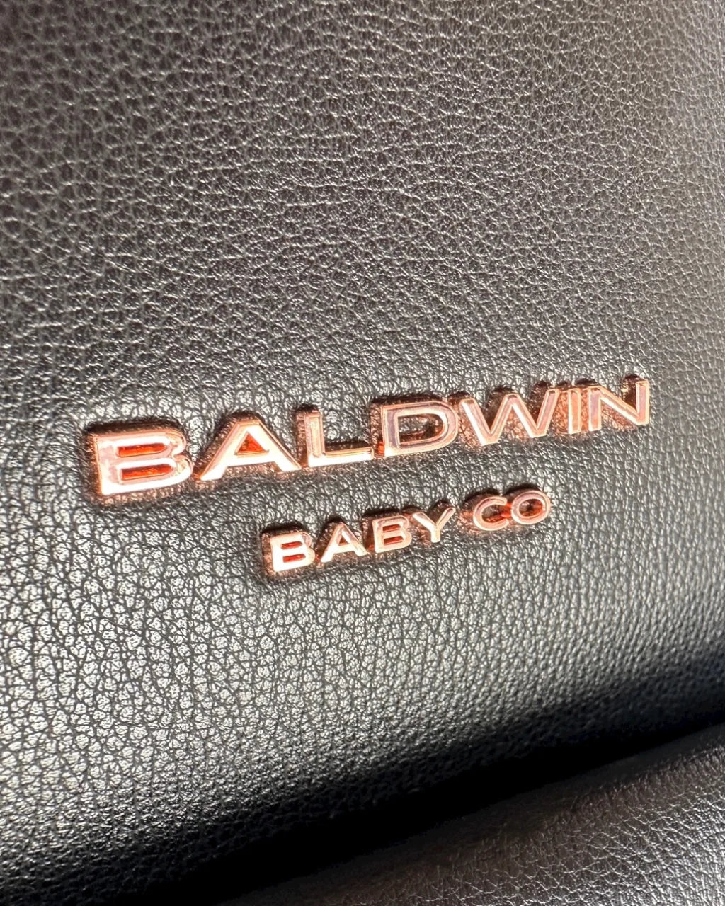 Close-up of the Baldwin Baby Co. logo in gold against the textured black vegan leather exterior of the diaper backpack.