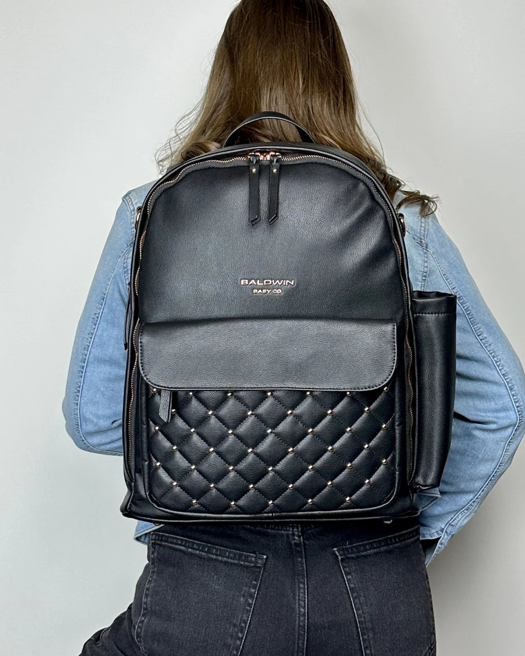A woman in a denim jacket and black denim wearing the black vegan leather diaper backpack on her back, showcasing its stylish and functional design for parents.