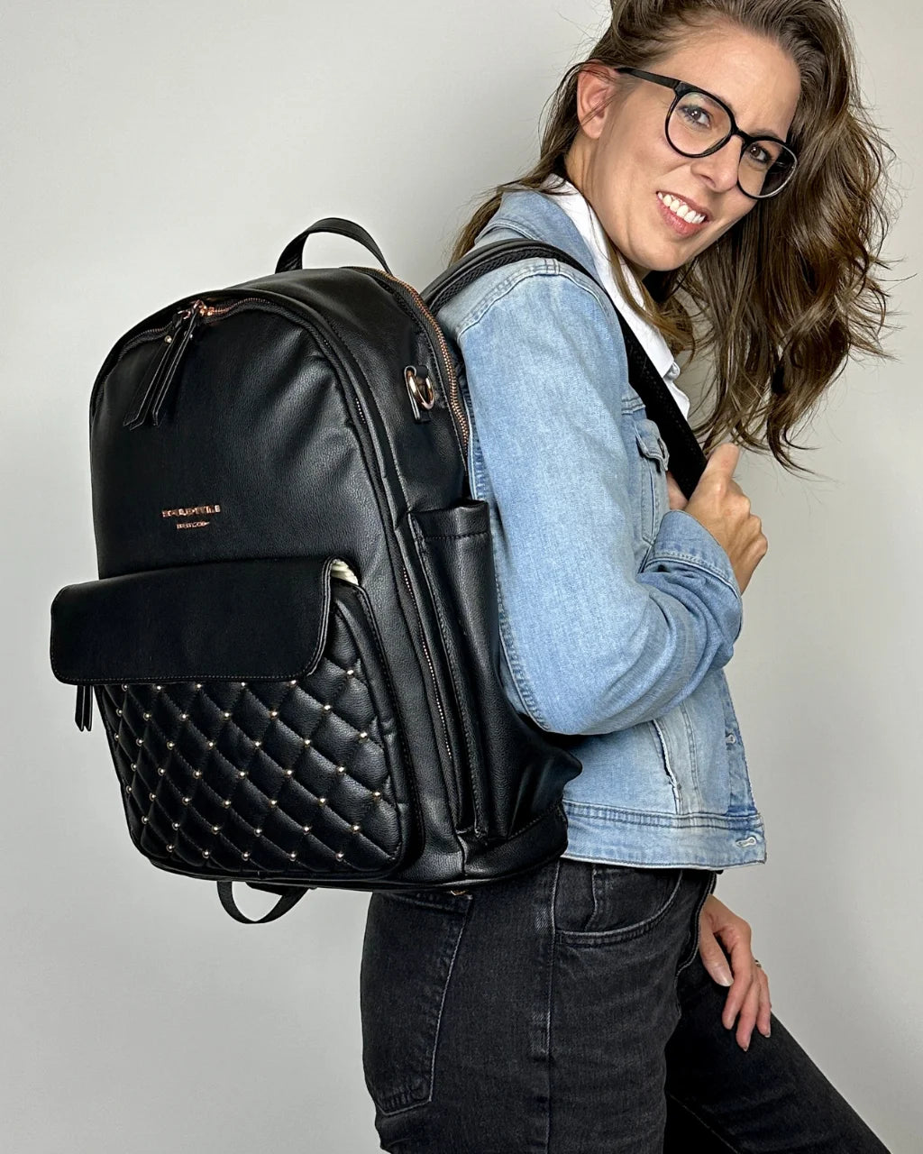 A smiling woman wearing the black vegan leather diaper backpack on her shoulder, highlighting its quilted pocket, red interior lining, gold hardware, and chic look.
