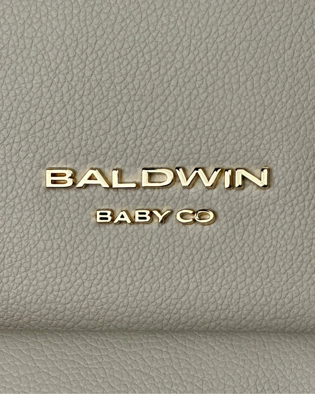 Close-up of the Baldwin Baby Co. logo in gold against the textured gray-beige vegan leather exterior of the diaper backpack.