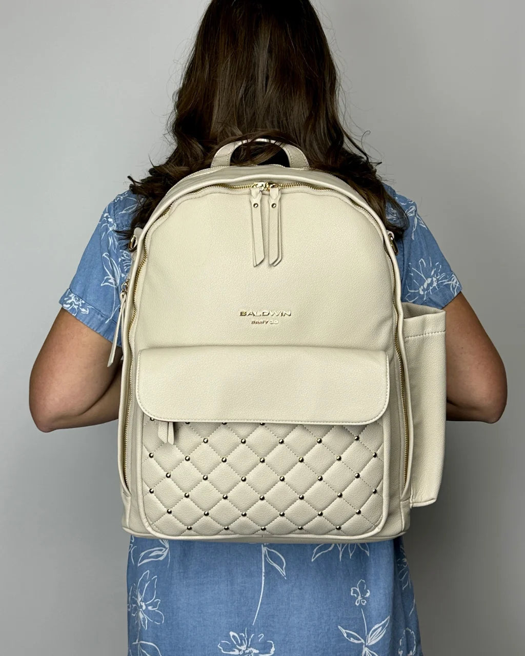 A woman in a blue dress wearing the gray-beige vegan leather diaper backpack on her back, showcasing its stylish and functional design for parents.