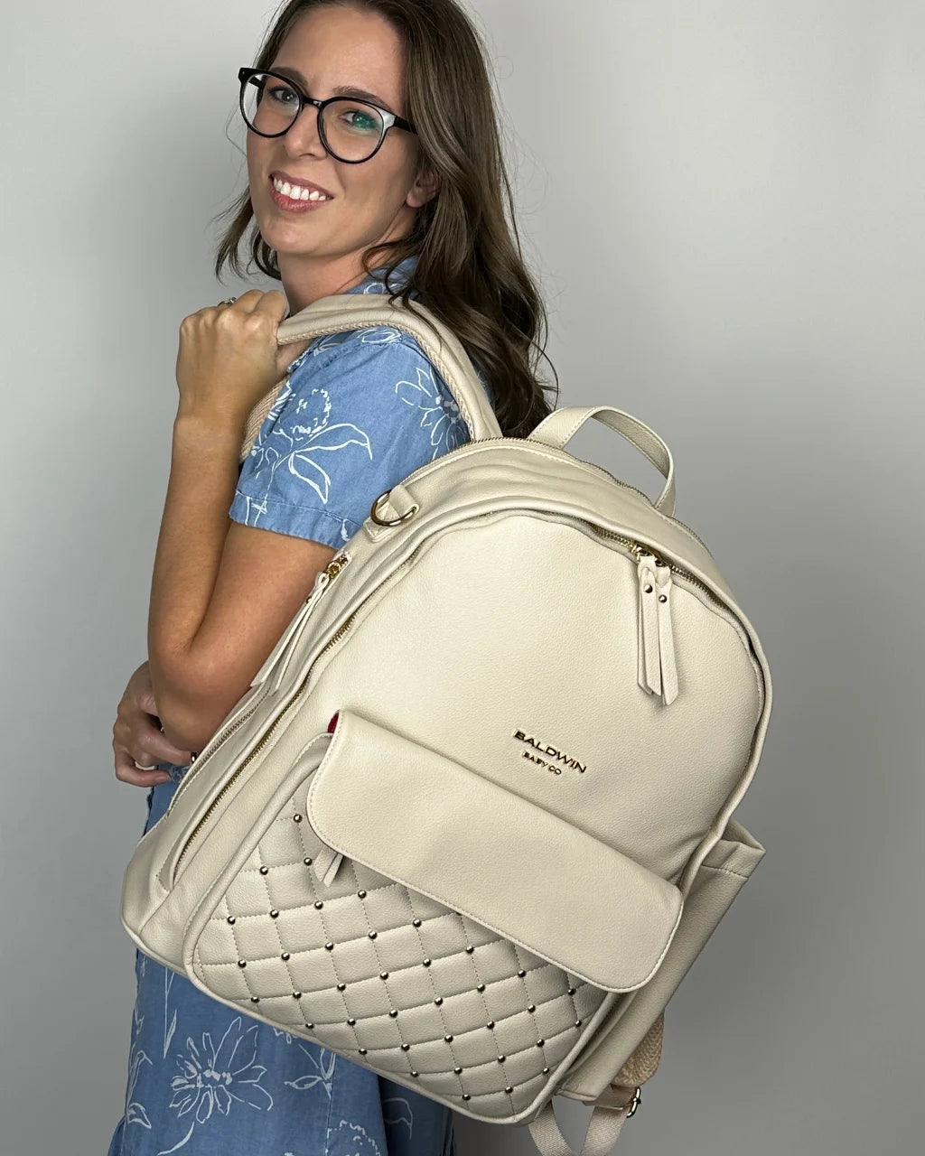 A smiling woman wearing the gray-beige vegan leather diaper backpack on her shoulder, highlighting its quilted pocket, red interior lining, gold hardware, and chic look.