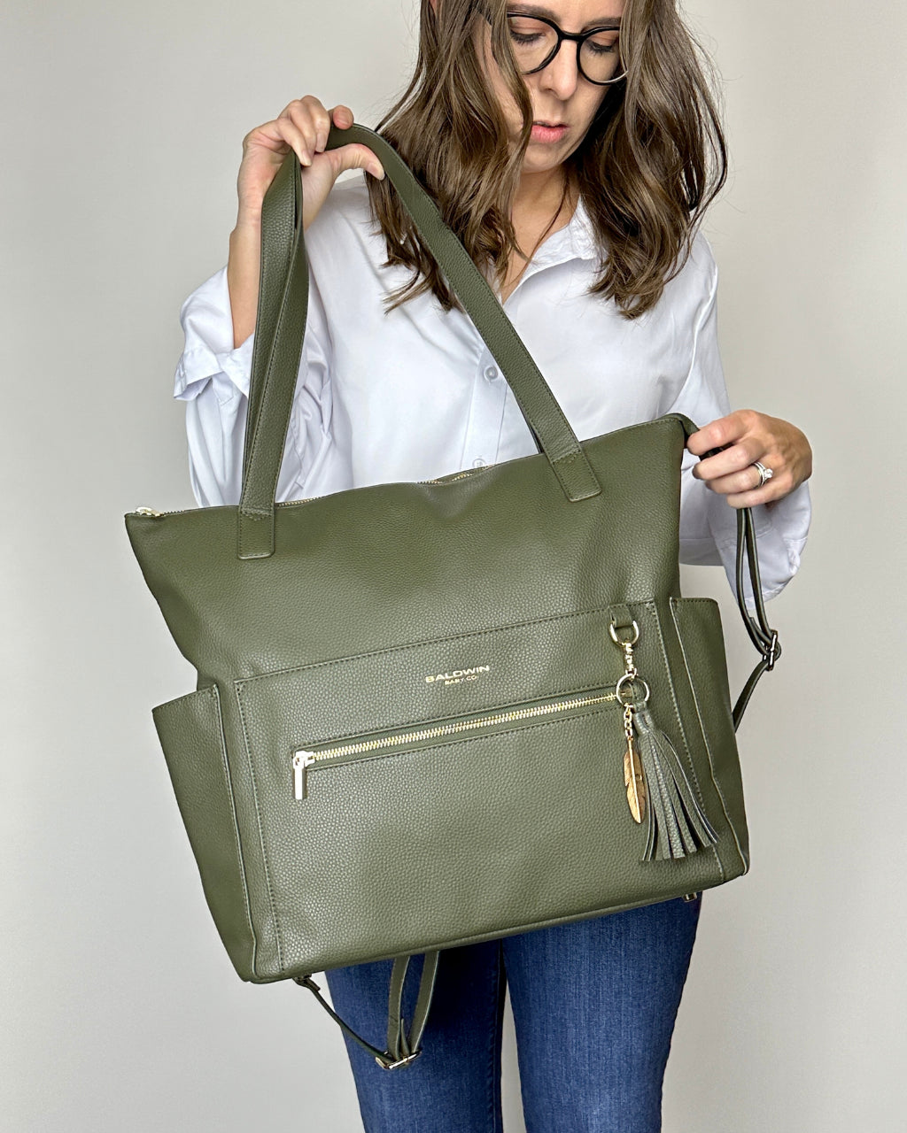 Woman holding green leather diaper bag with gold feather and tassel by the shoulder straps