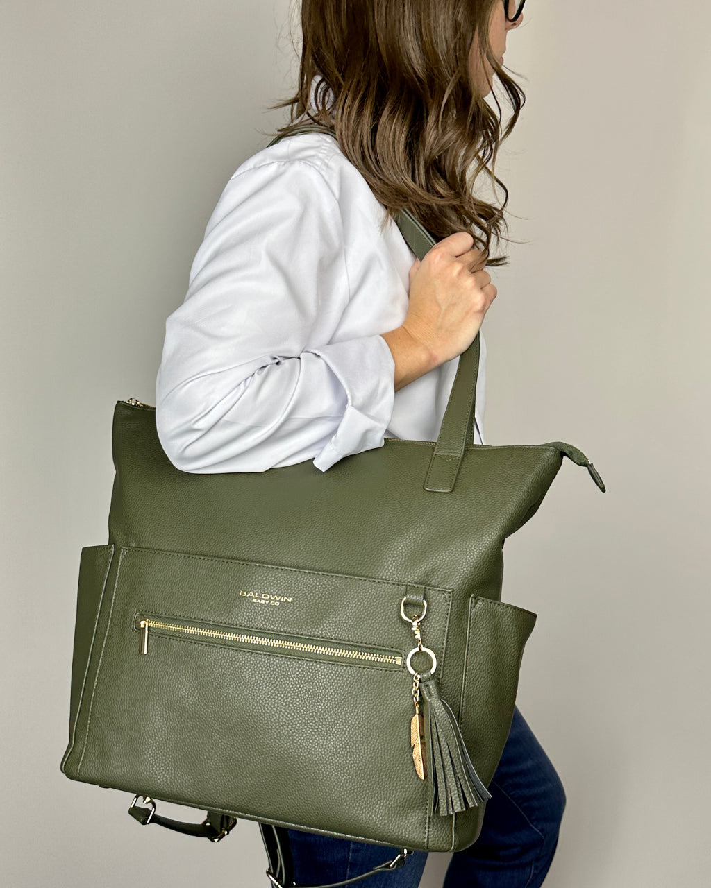 Woman holding green leather diaper bag tote with gold feather and tassel on her should by the shoulder straps