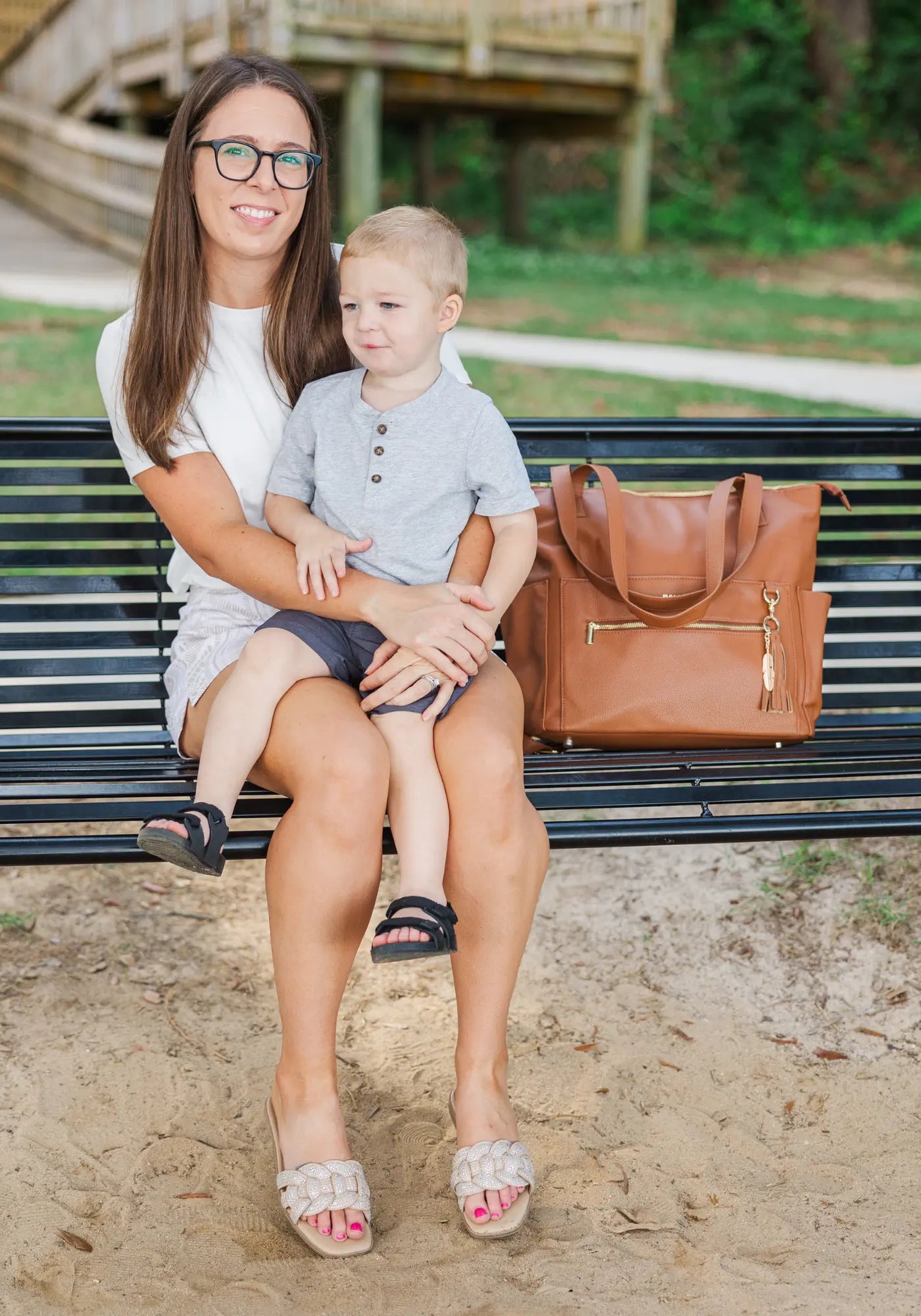 Woman with glasses holding a young boy on a park bench, brown diaper bag beside them.