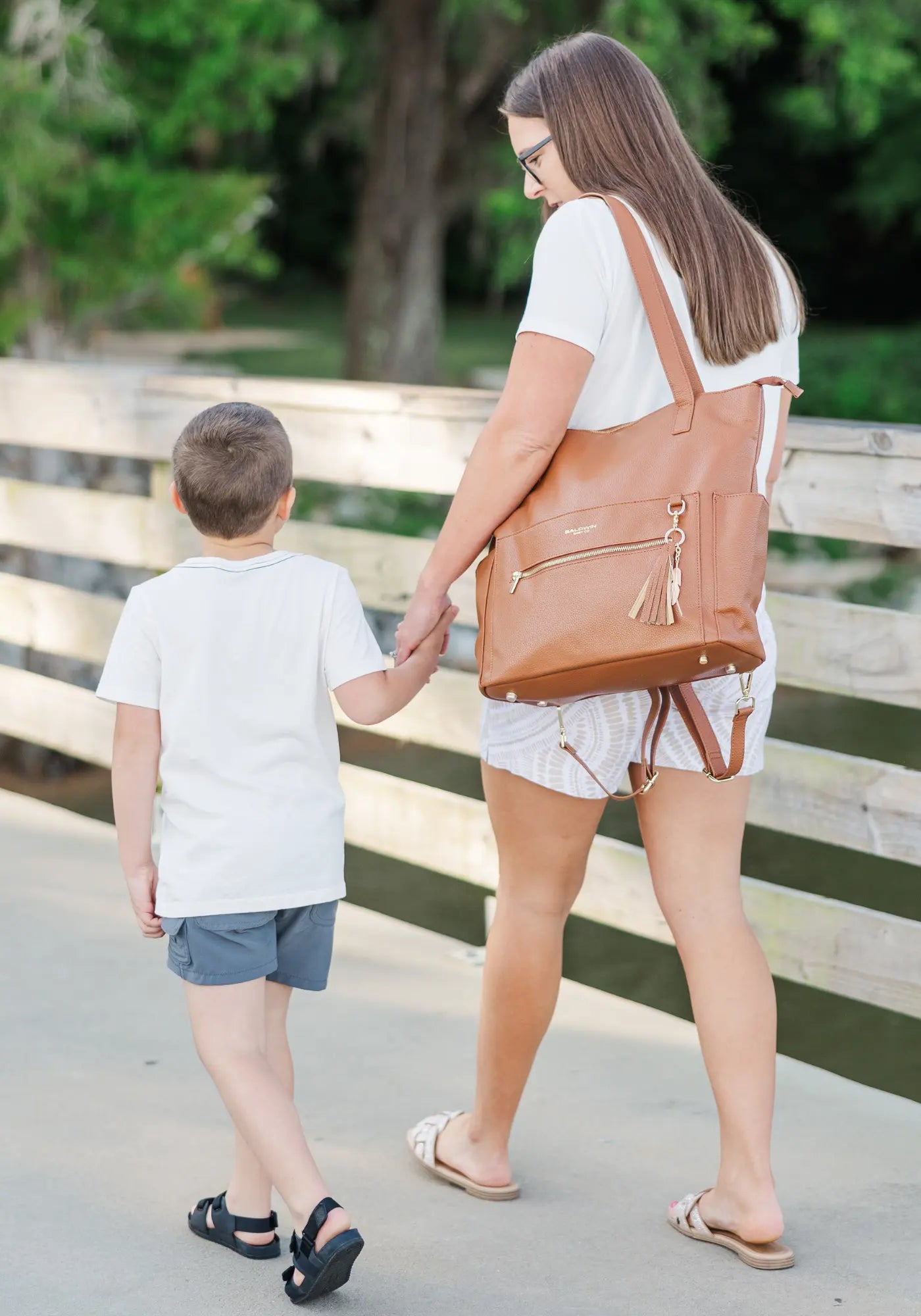 Woman with brown diaper bag holding hands with a young boy on a wooden walkway outdoors.