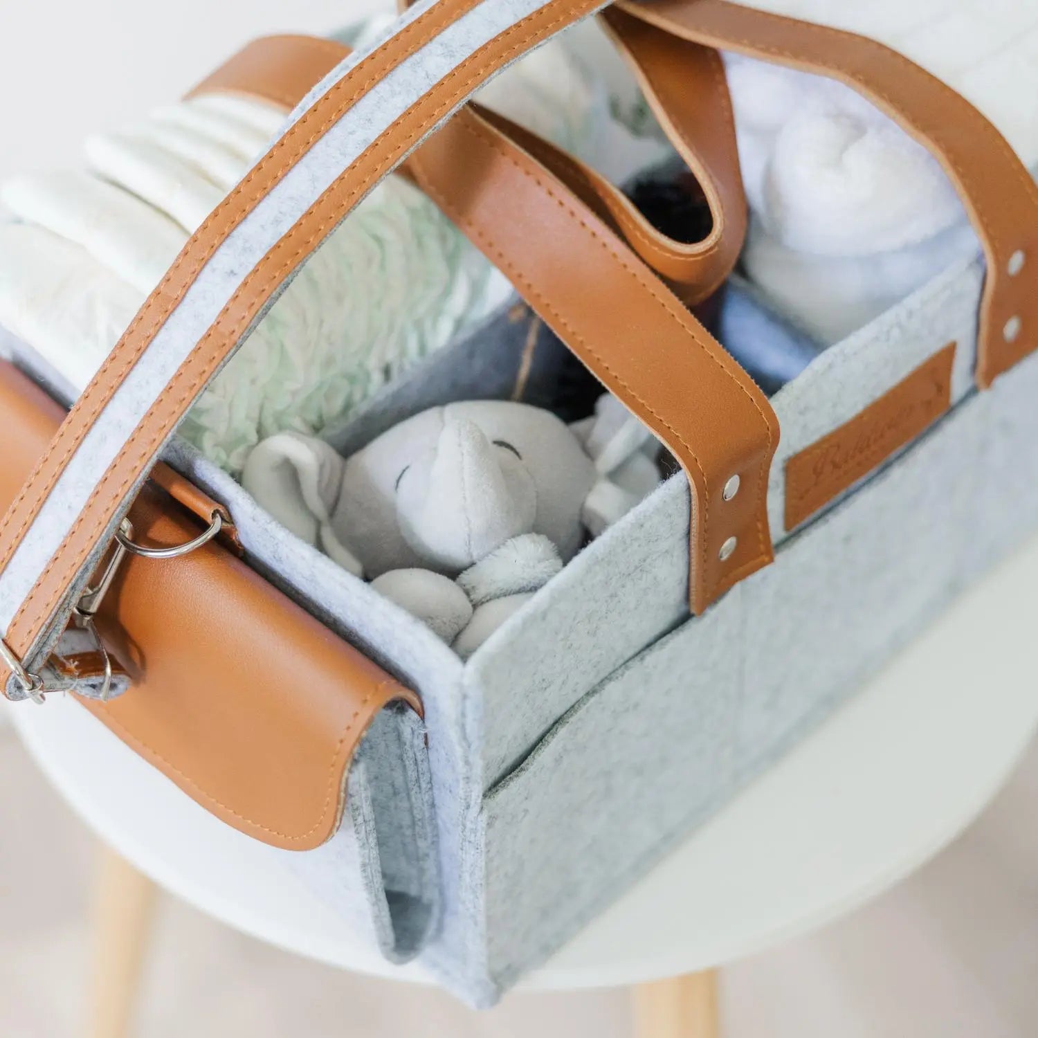 Gray felt diaper caddy with brown leather handles and baby items on wooden table