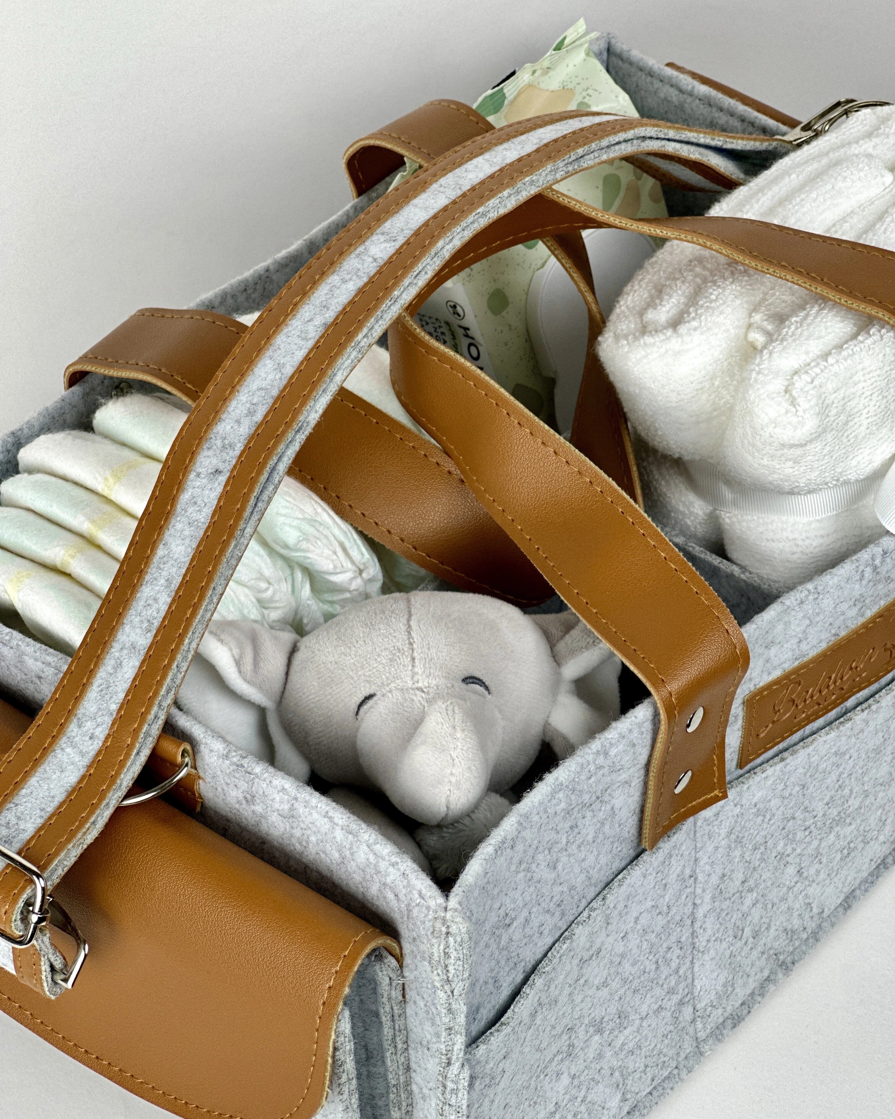Gray felt diaper caddy with brown leather handles, filled with baby items, on a wooden table.