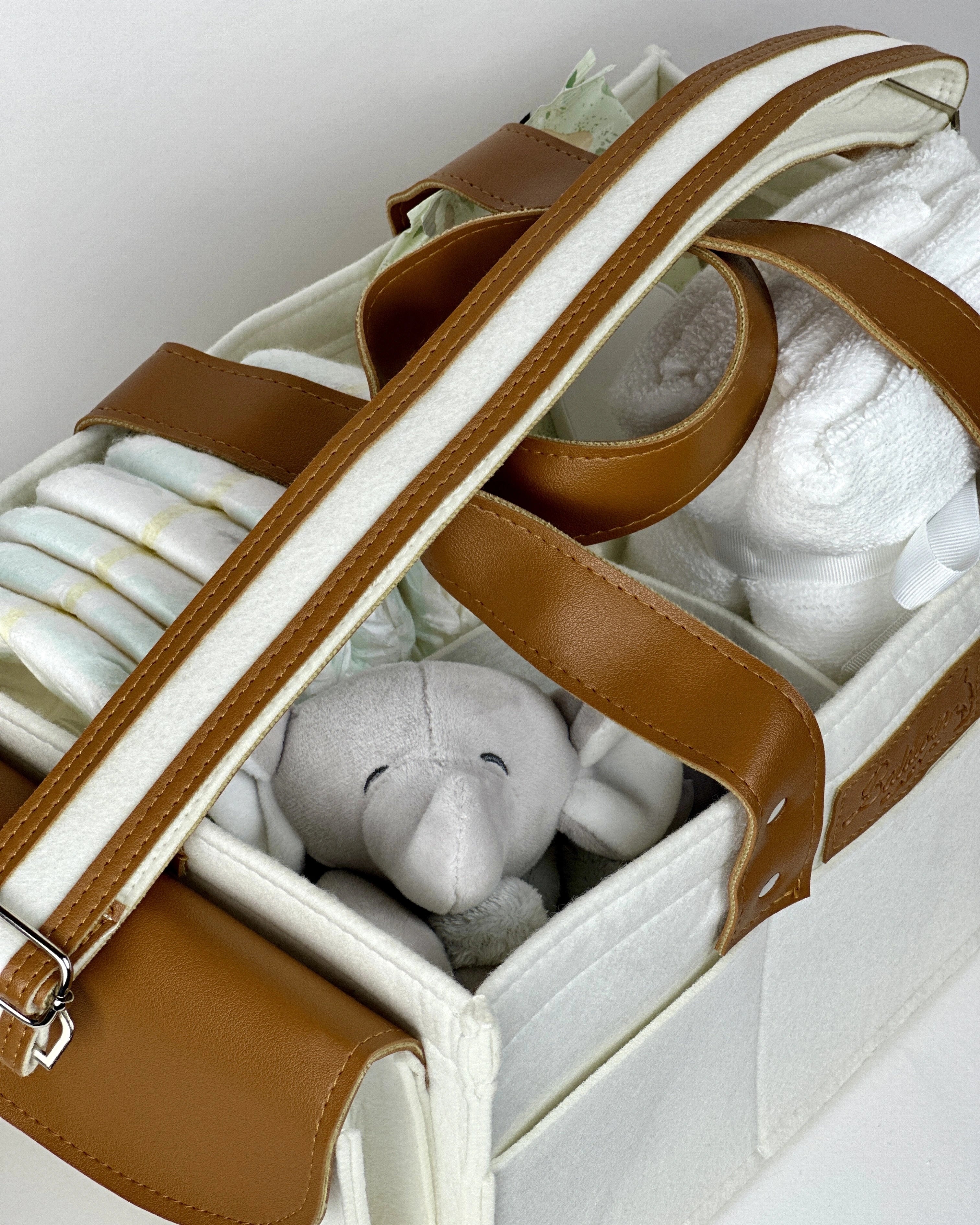 White felt diaper caddy with brown leather handles, filled with baby items, on a wooden table.