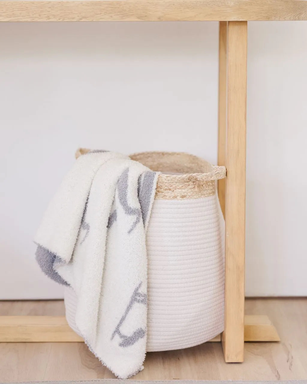 white blanket with gray herons draped over a white bell shaped rope basket with corn husk details and handlesr
