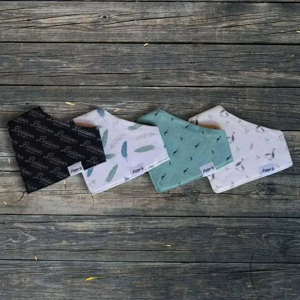 Set of four bandana bibs on a wood background. Charcoal with light gray Baldwin Bay Co pattern, white with green and transparent feathers, green with dark gray herons, and creamy white with Baldwin Baby Co's signature heron pattern