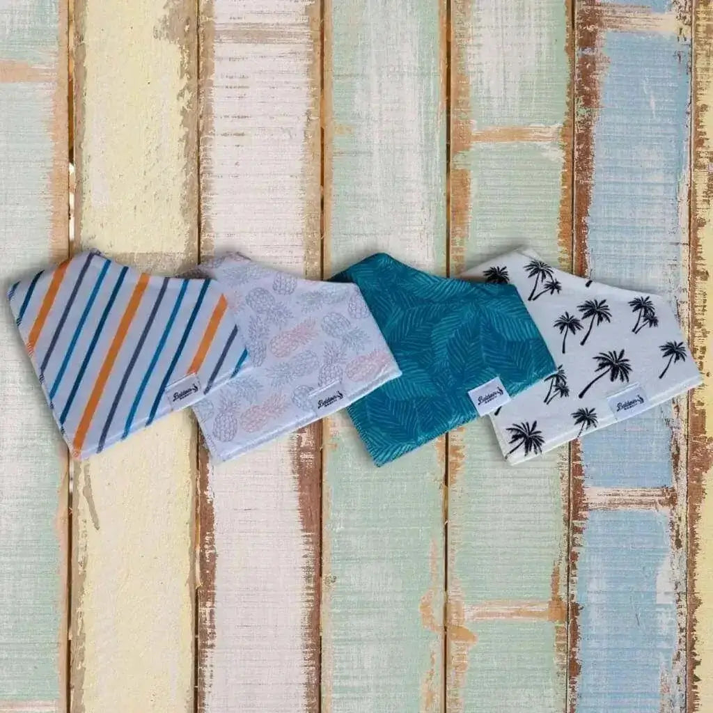 Set of four bandana bibs on a wooden pastel background. White, blue, gray, and orange stripes; white with blue, pink, and green pineapples; green monstera; and creamy white black palm tree patterns
