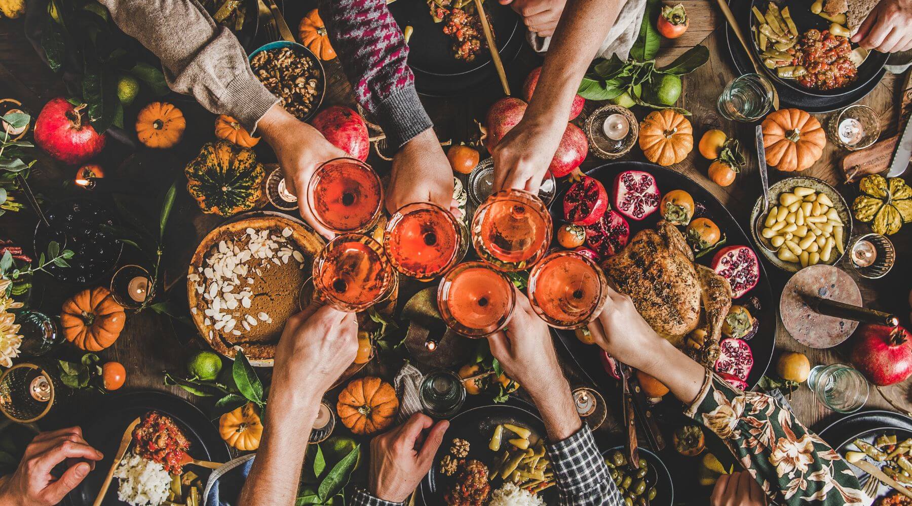 A top view of a Thanksgiving feast where friends and family toast with glasses of rosé around a table full of traditional holiday dishes, including pumpkin pie, roasted turkey, pomegranate seeds, and small decorative pumpkins.