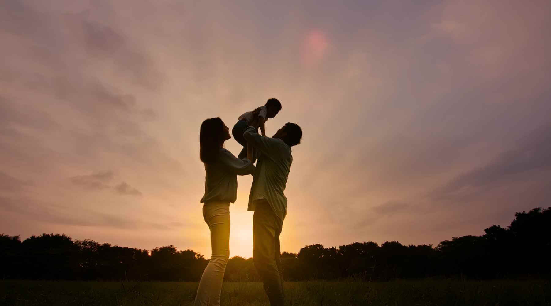 A silhouette of a family outdoors at sunset, with a mother and father lifting their baby in the air against a colorful sky.