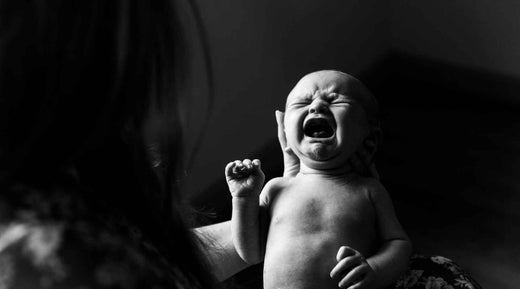 A black-and-white close-up of a newborn baby crying while being gently cradled by an adult's hands.