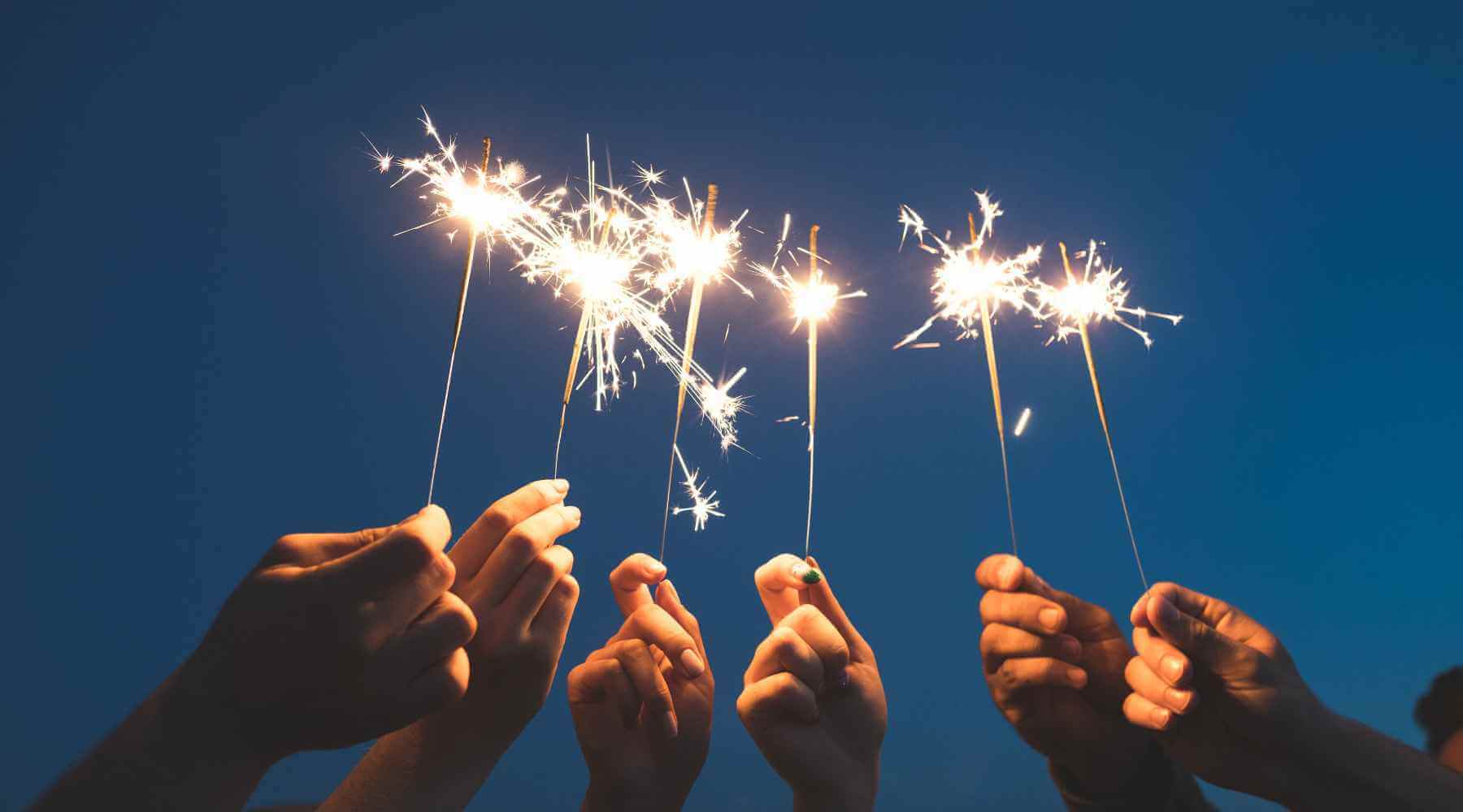 Several hands holding lit sparklers against a dark sky, capturing the excitement and celebration of ringing in the New Year with a bright and joyful sparkler display.