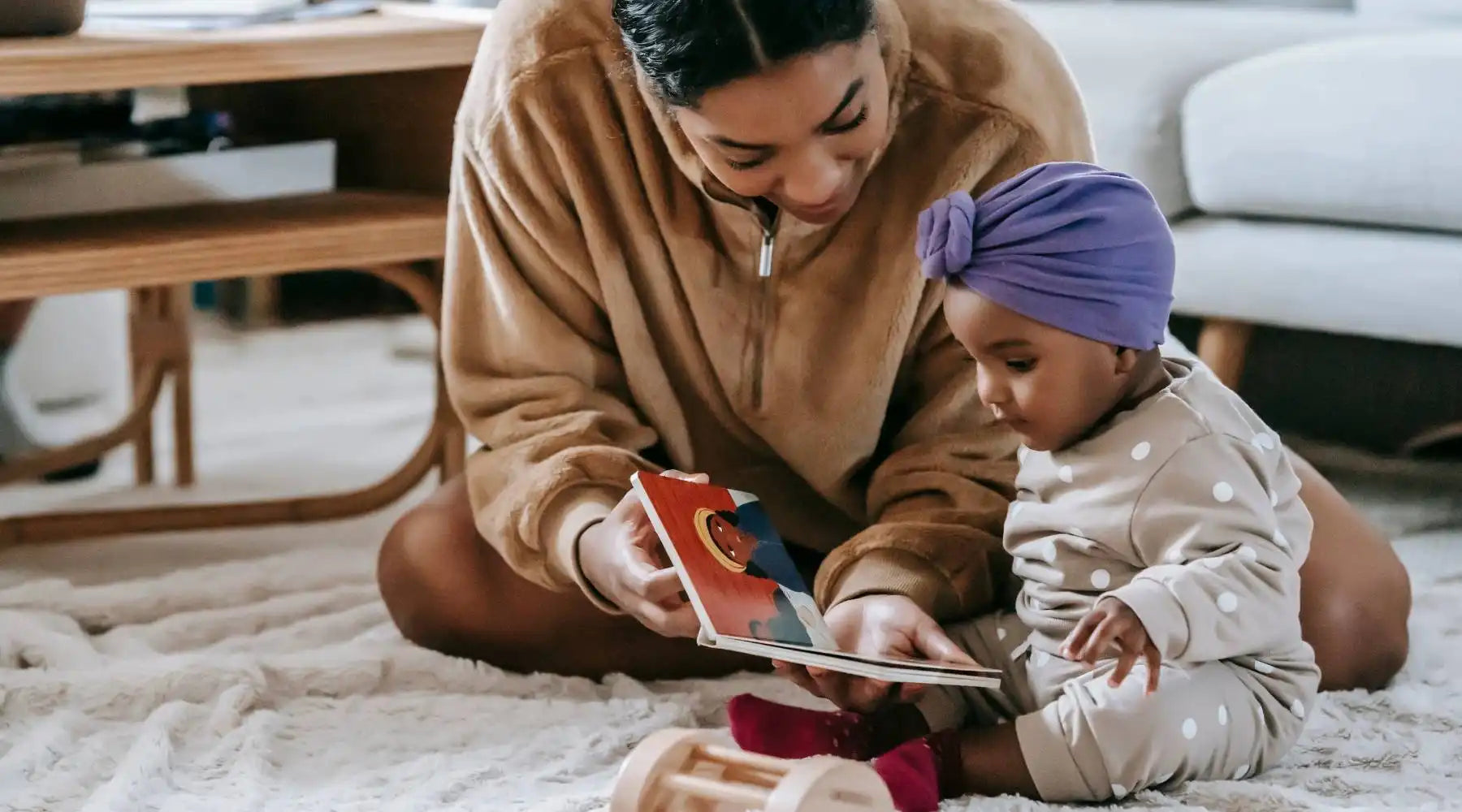 Mother reading colorful board book to baby girl wearing purple headwrap, nurturing early literacy skills.