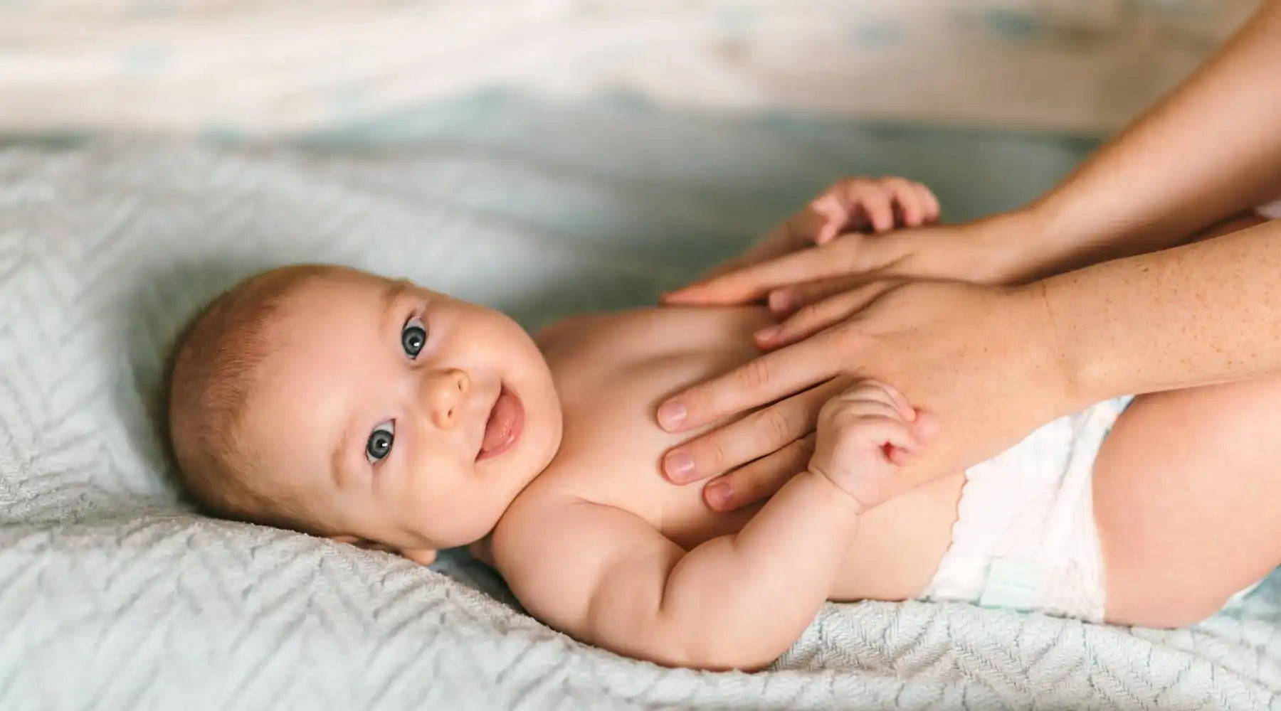 Smiling baby in diaper enjoys gentle tummy massage on soft blanket, highlighting baby skincare moments.