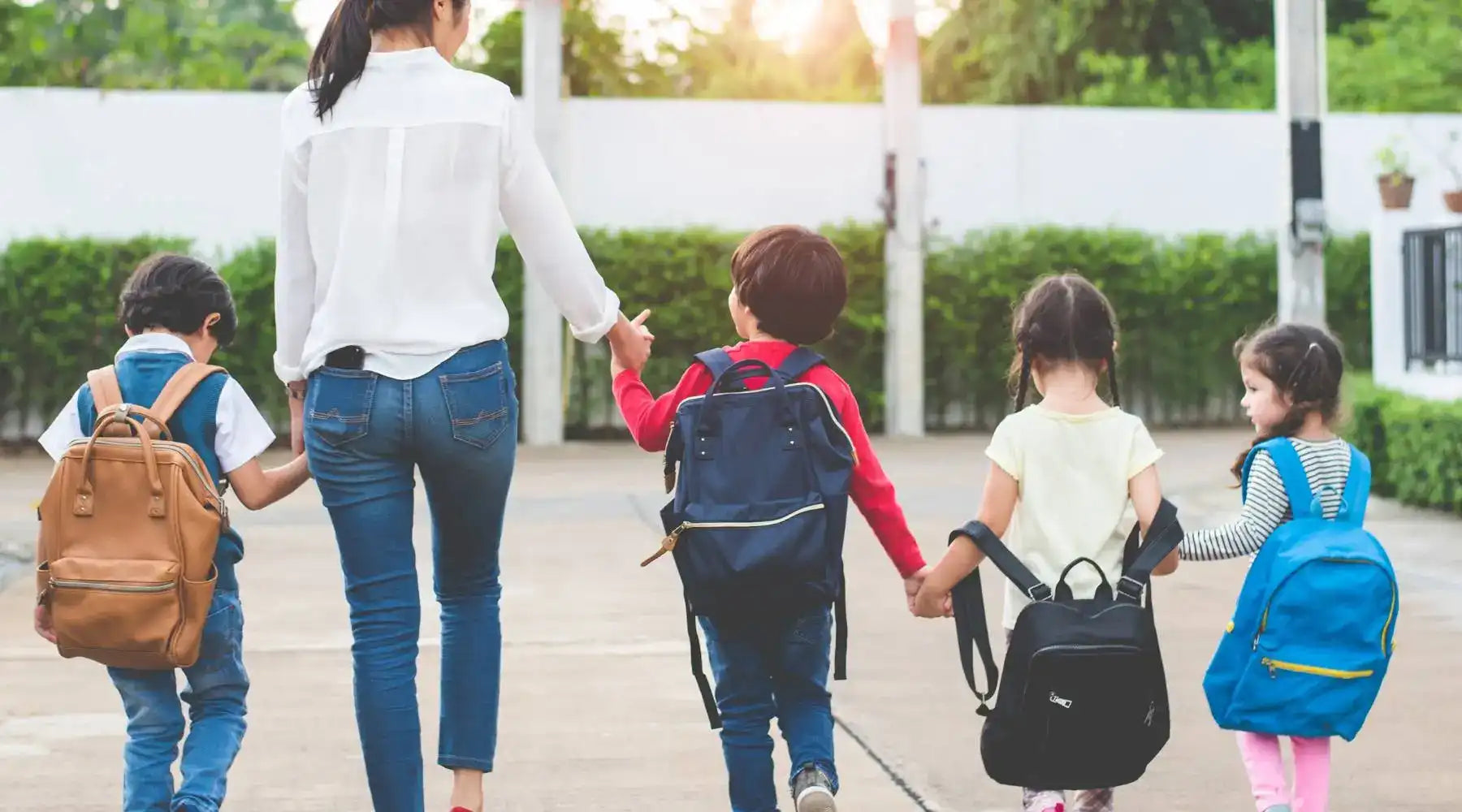 Mother holds hands with four young children wearing colorful backpacks on the way to school.