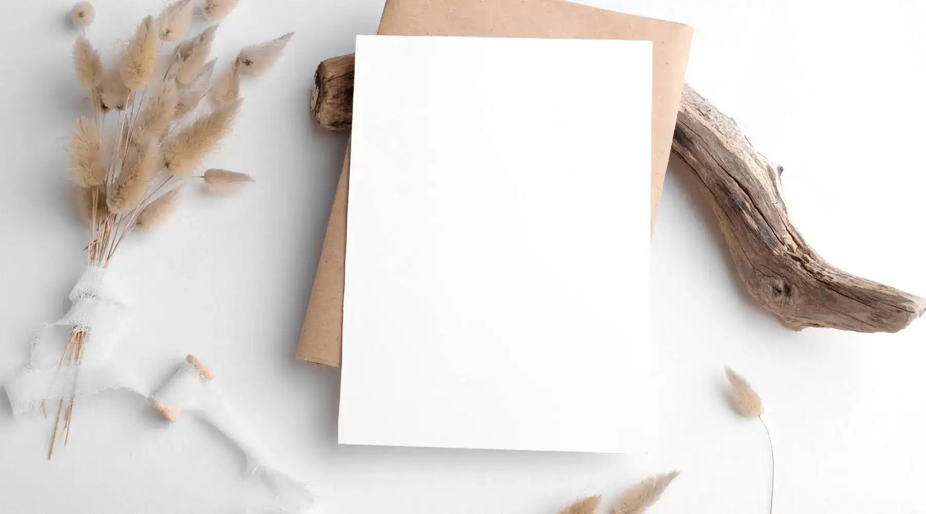 Minimalist flat-lay with blank white card, dried florals, kraft paper, and driftwood on a white background.