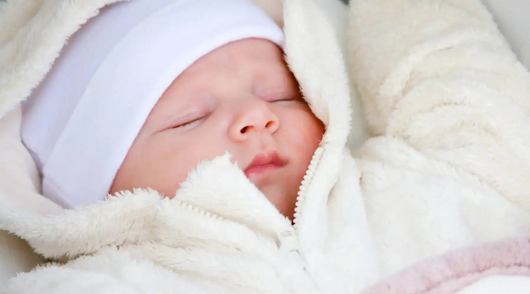 Newborn baby peacefully sleeping, dressed in a white fuzzy outfit and white hat.