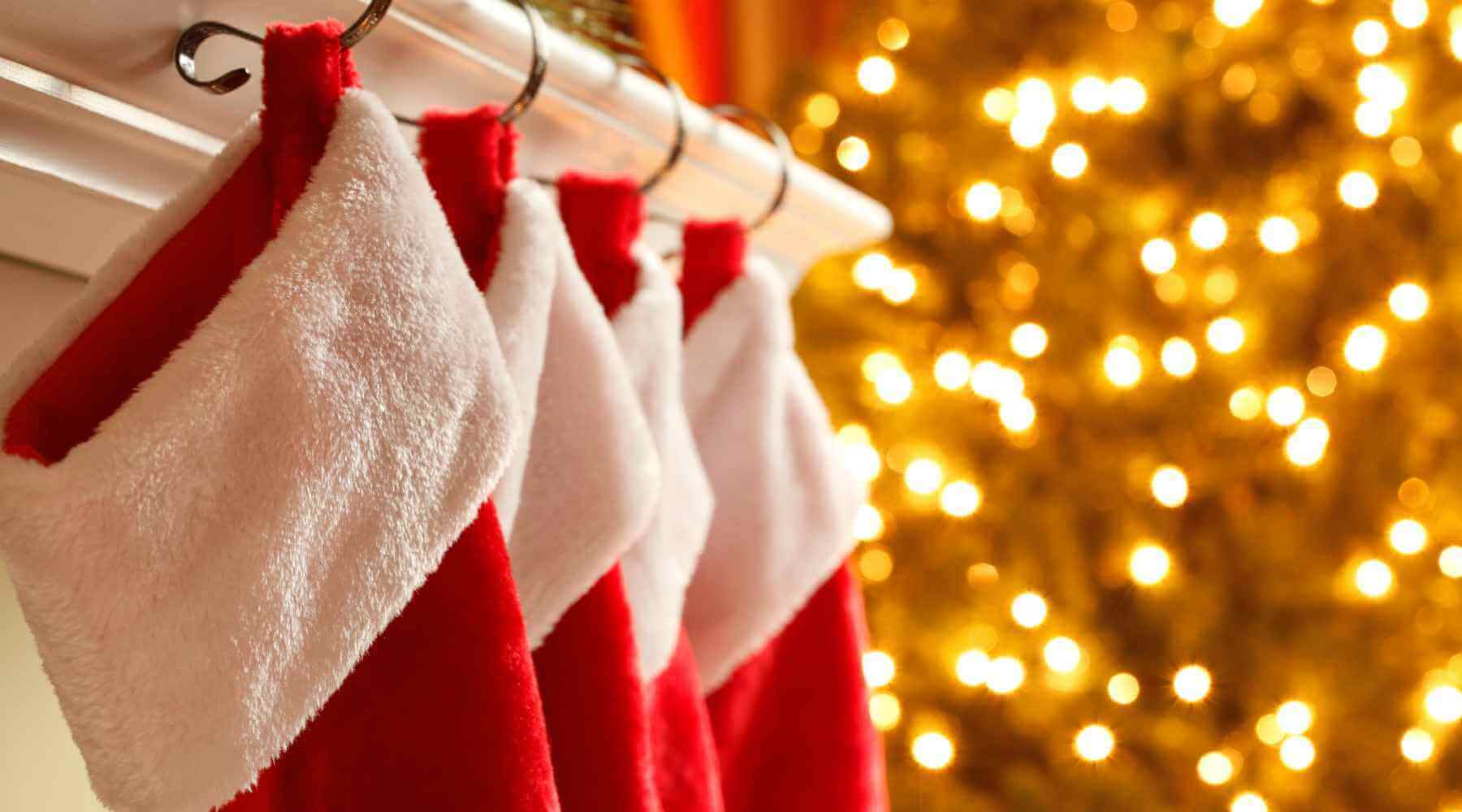 Four red and white Christmas stockings hanging from hooks on a mantel, with a beautifully lit Christmas tree glowing softly in the background.