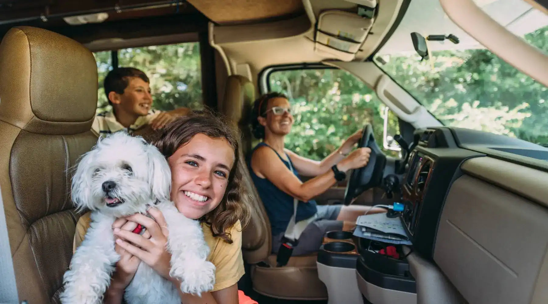 Smiling family traveling inside an RV, with a girl holding a fluffy white dog in the front seat while mom drives.
