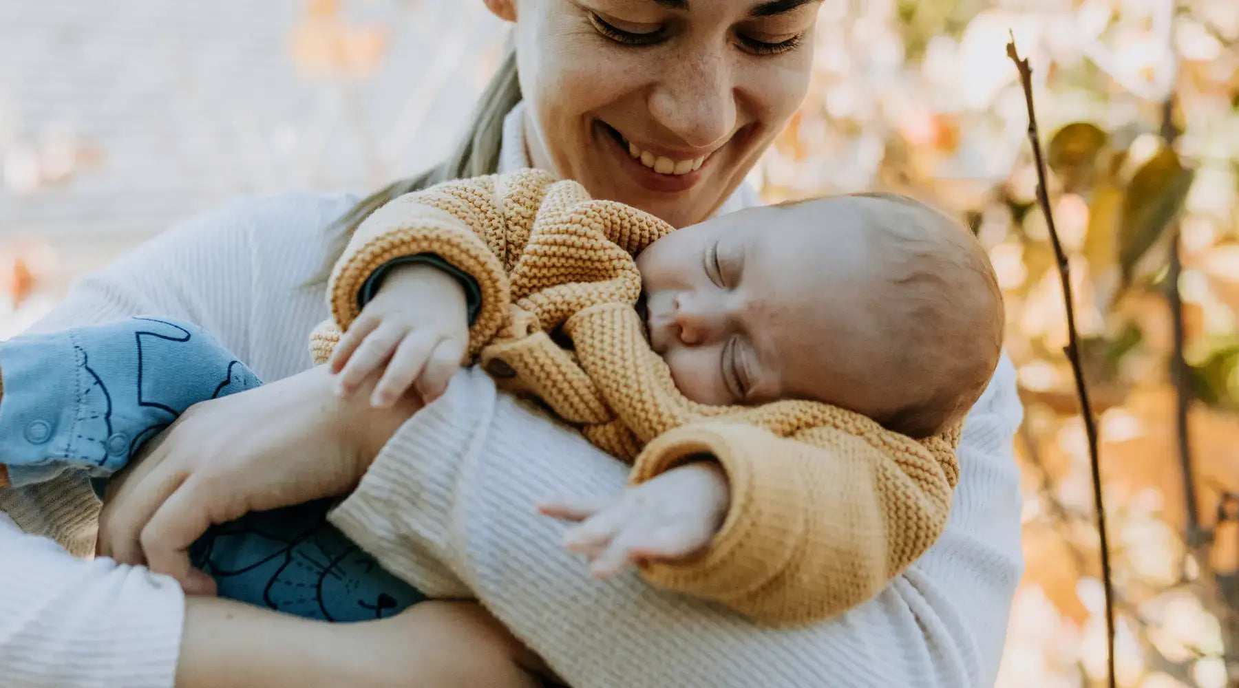 A mother in a cozy sweater smiles as she cradles her sleeping baby in her arms. The autumn leaves in the background add warmth to the scene, reflecting a peaceful and joyful motherhood moment.