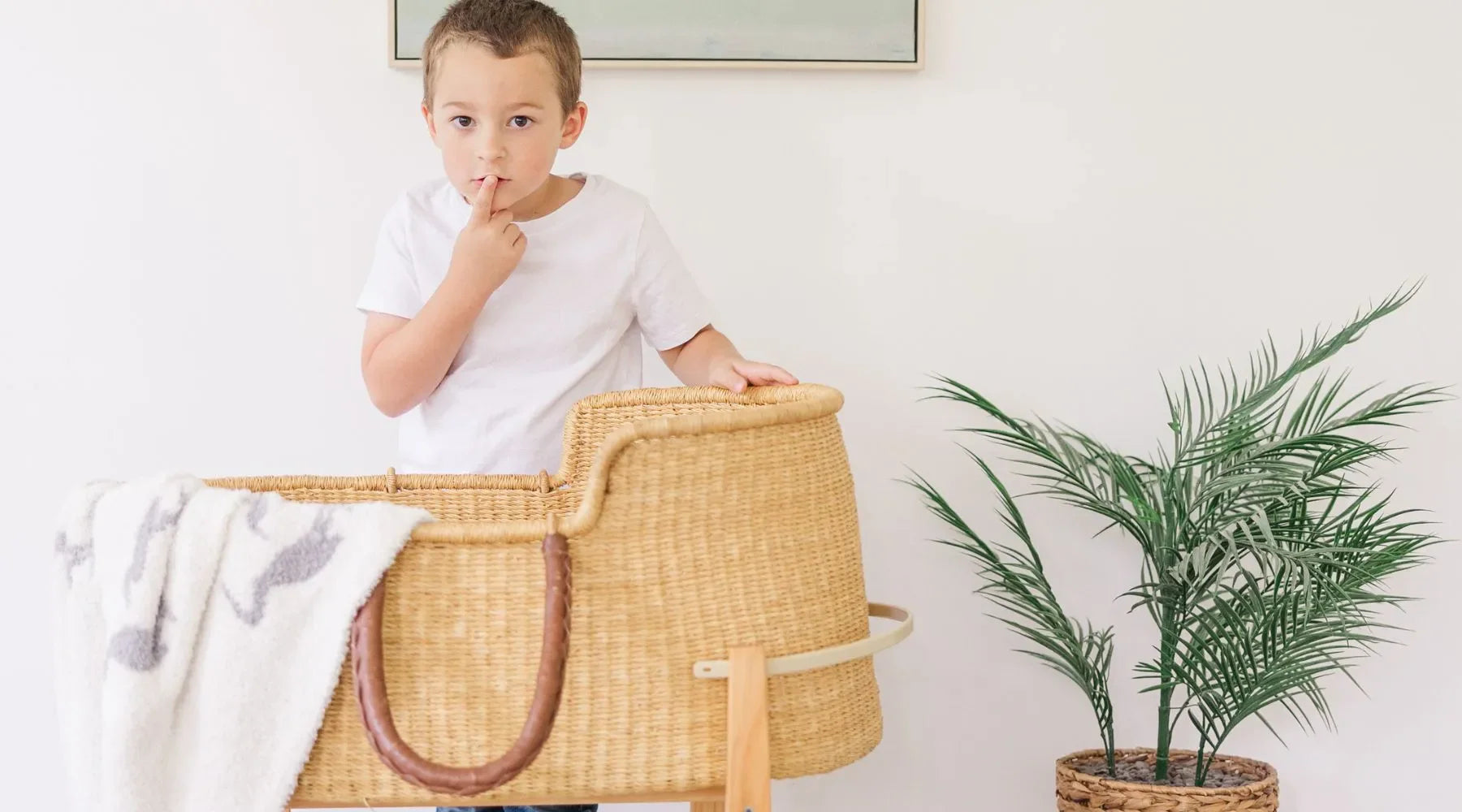 A young boy standing next to a wicker bassinet, looking curious and making a "shh" gesture with his finger.