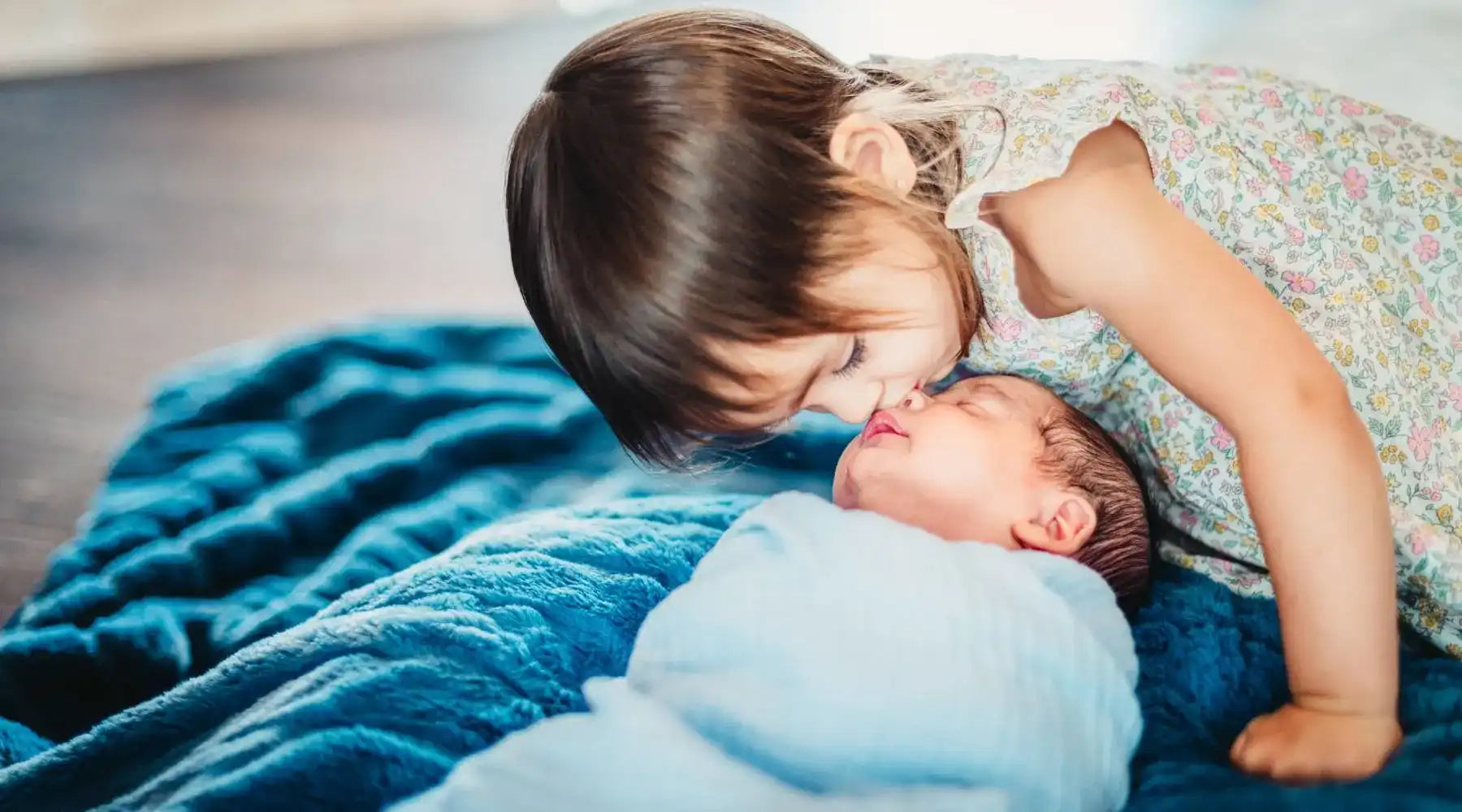 Little girl in a floral dress leaning over to kiss a newborn baby swaddled in a soft blue blanket.